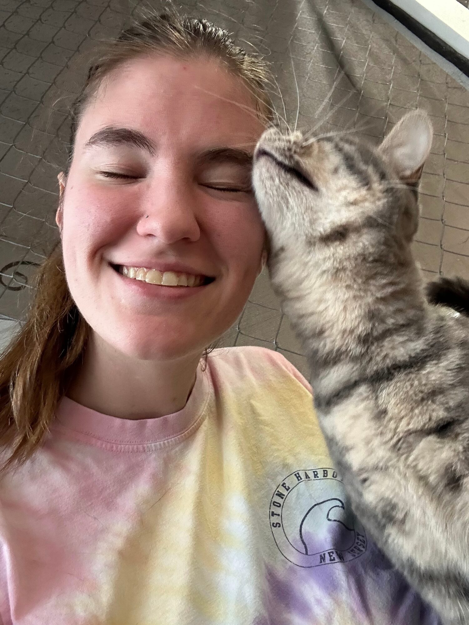 Person smiling with eyes closed, a cat is licking their face, background shows a brick wall.