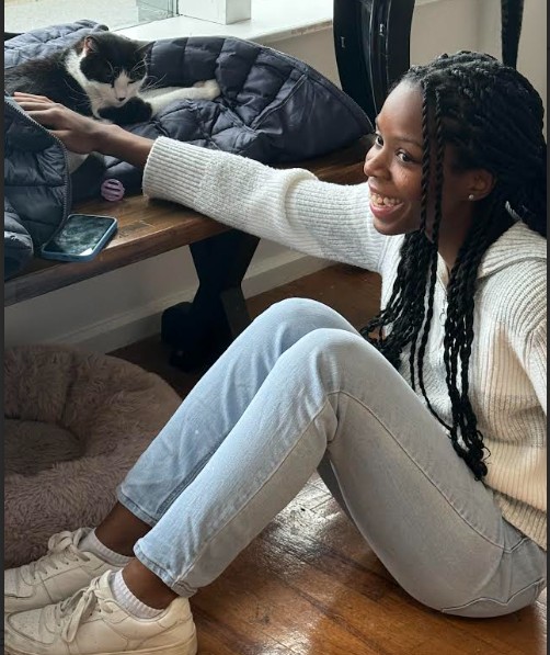 Girl with braided hair reaching to pet a black and white cat on a bed, smiling, sitting on the floor.