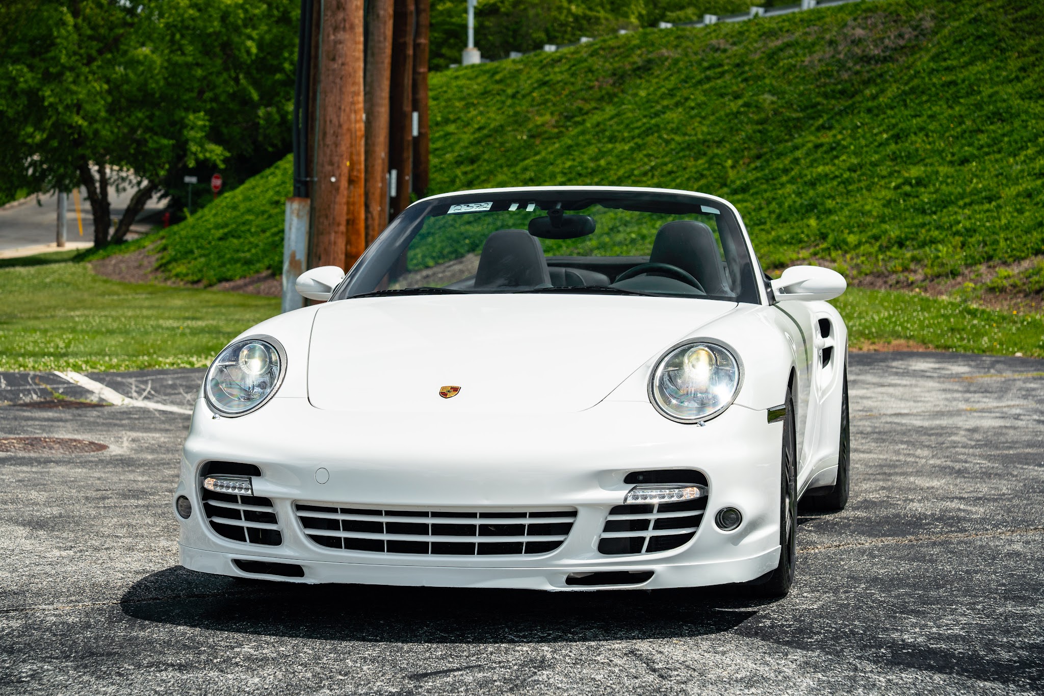 White convertible sports car parked on asphalt with green grass and trees in background.