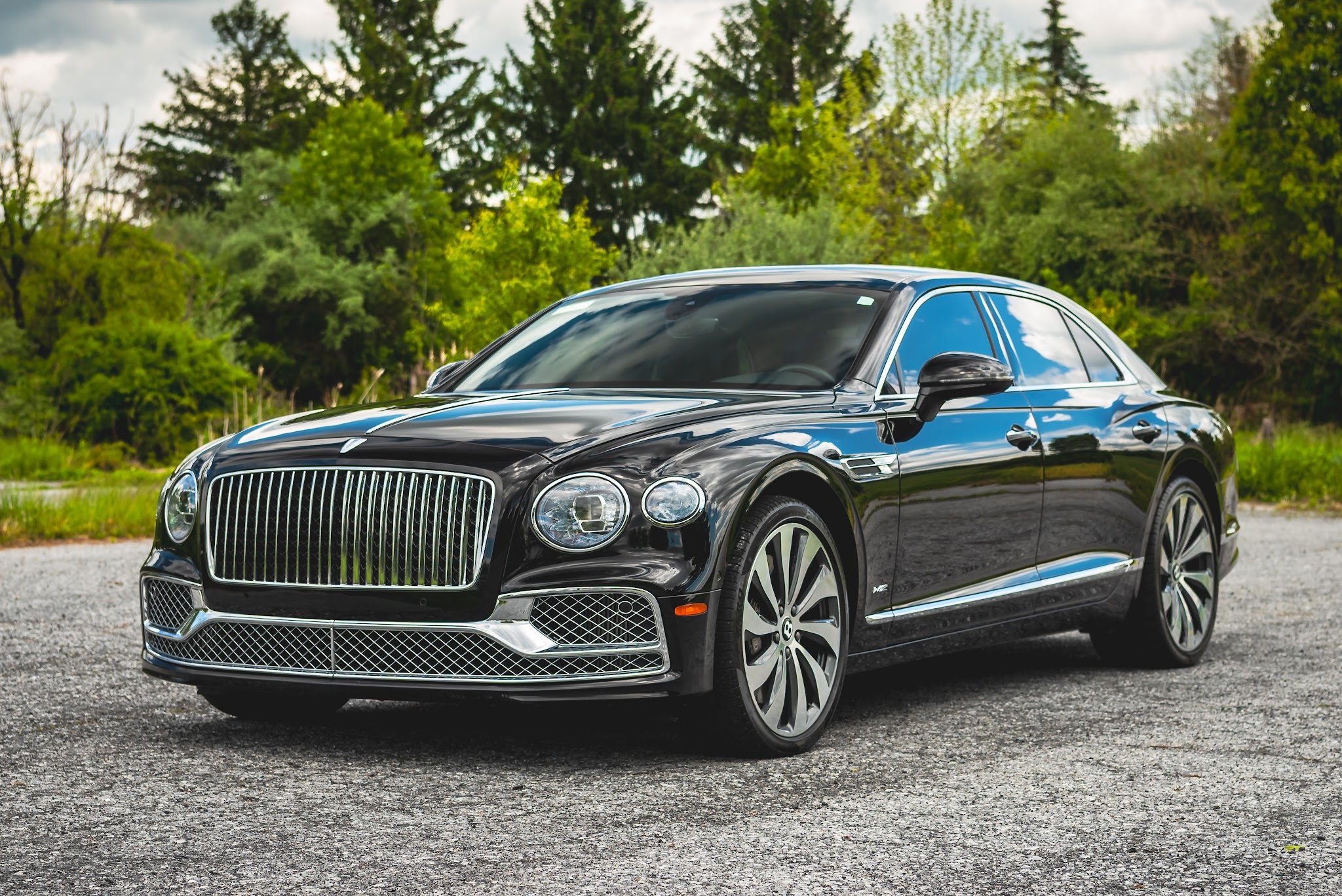 Black luxury sedan parked on a gravel surface with trees and greenery in the background.