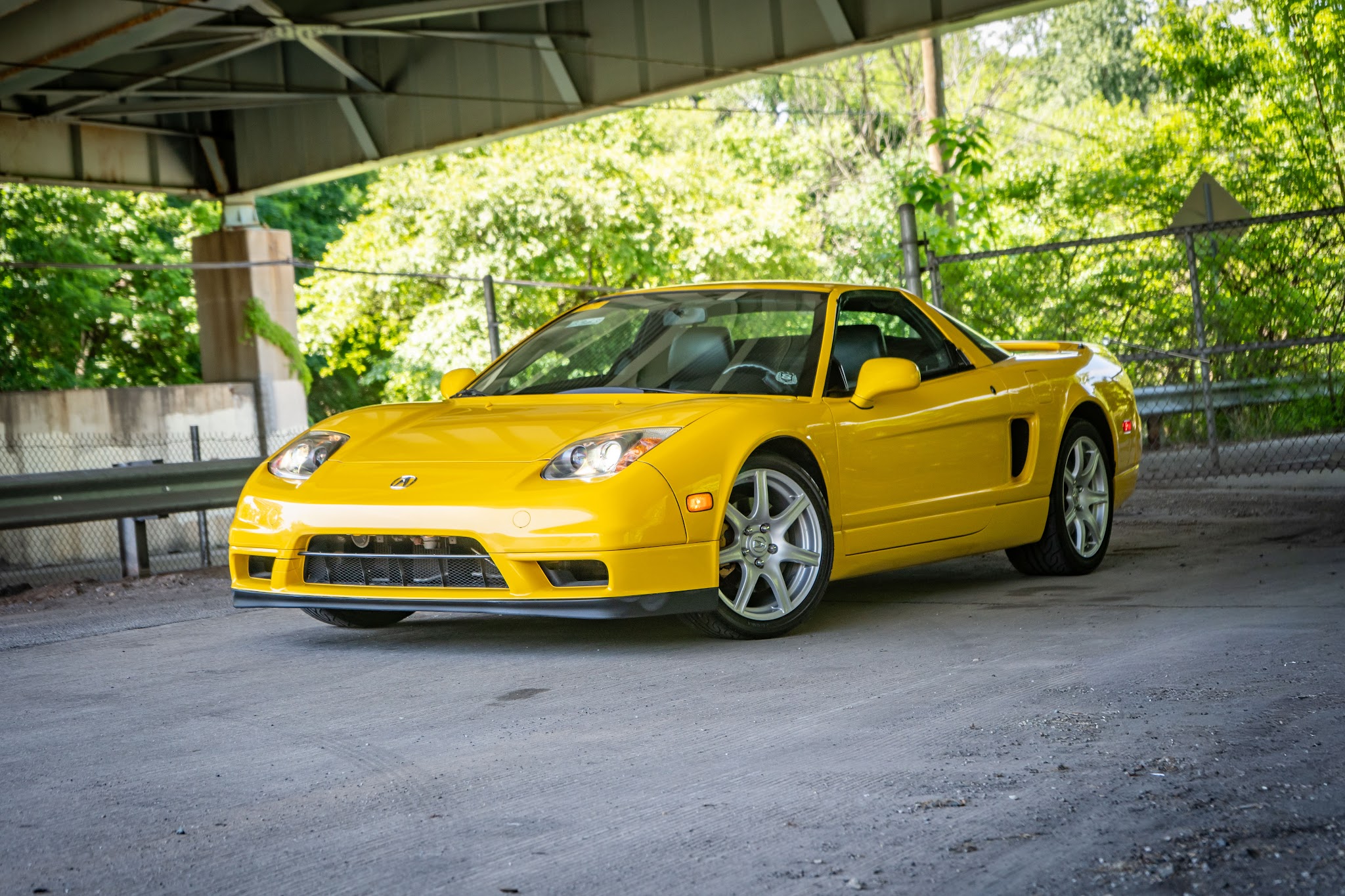 Yellow sports car parked under a bridge with green trees in the background.