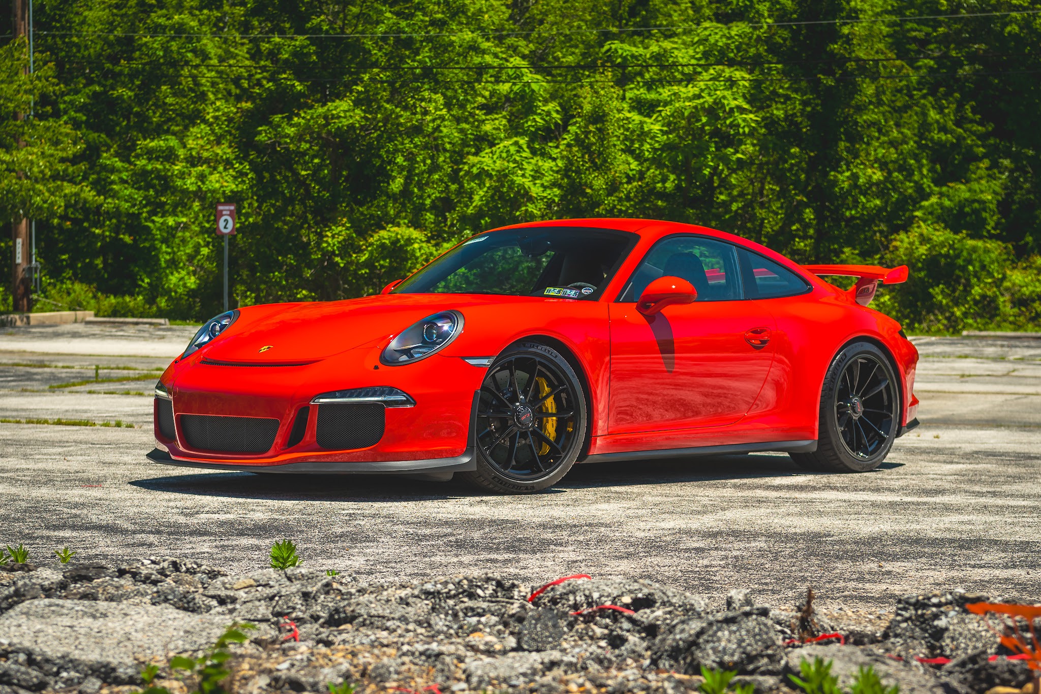 Red sports car parked on a paved surface with green trees in the background.