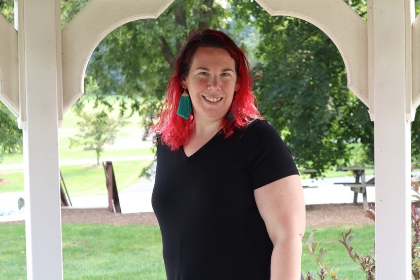 Woman with red hair smiling, wearing a black shirt and teal earrings, standing outdoors under a white archway.
