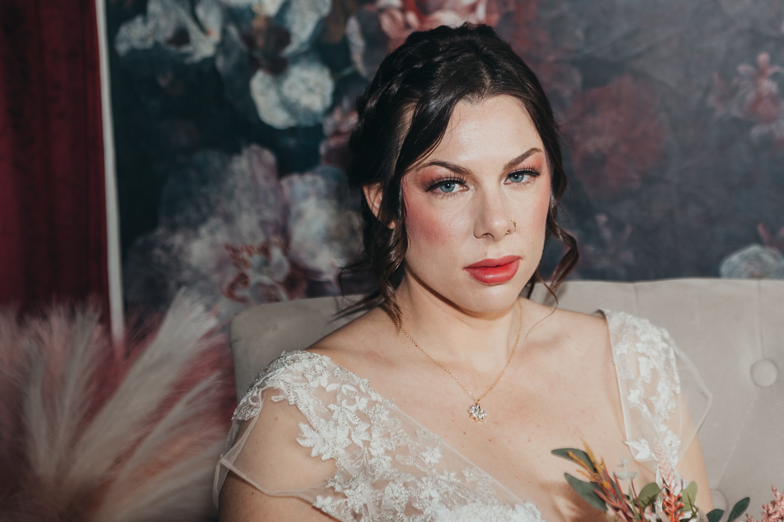 A woman with dark hair and makeup, wearing a white lace dress, sitting on a beige sofa with a floral background.