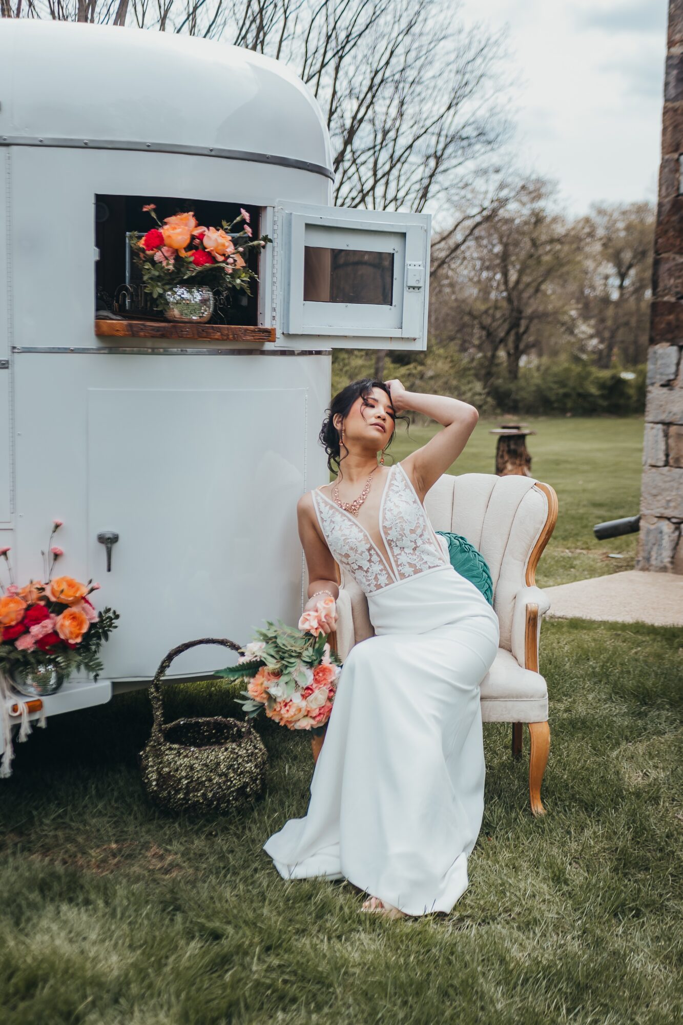 Woman in wedding dress sitting on a chair outdoors near a vintage trailer with flowers, hand on head, relaxed pose.