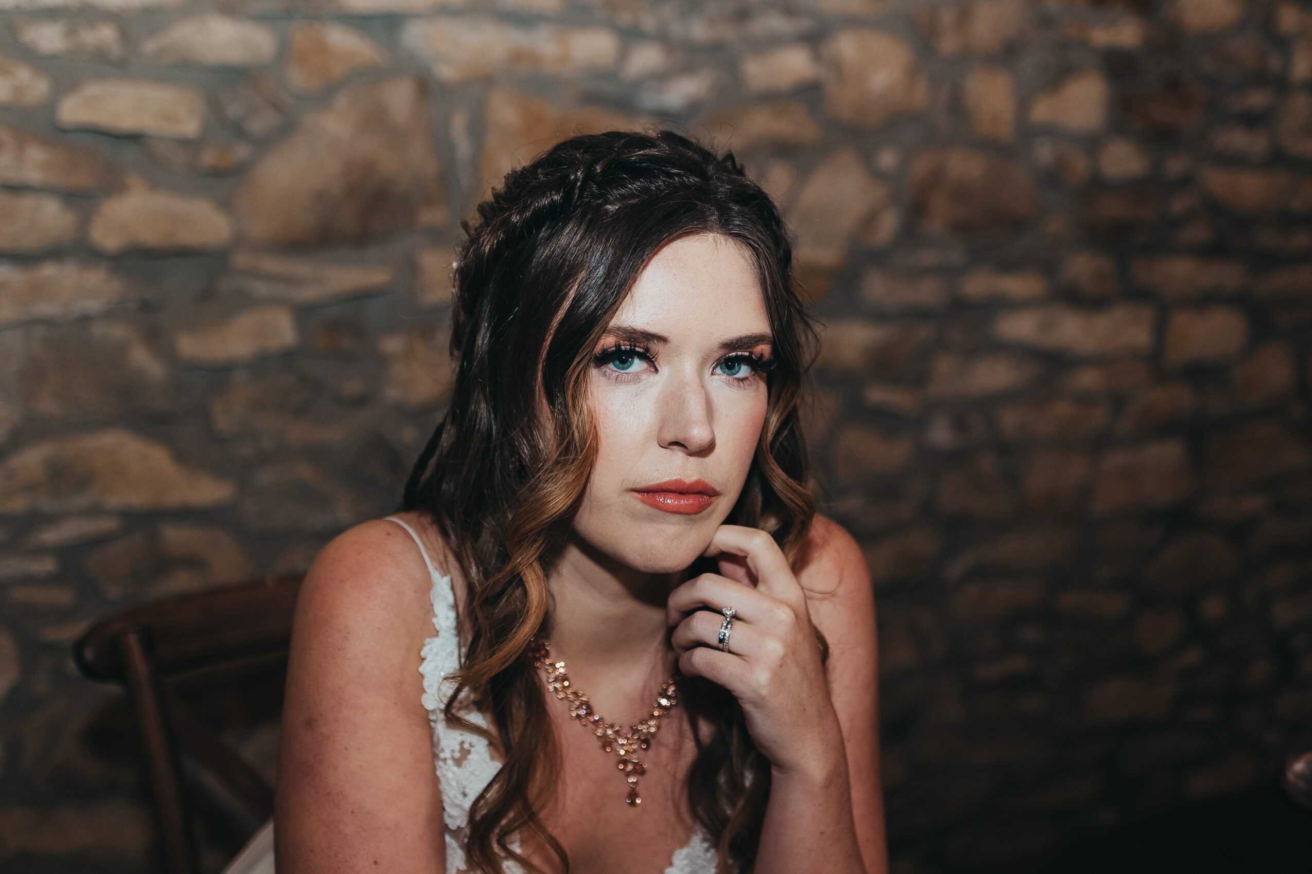 Woman with dark hair and makeup, wearing a necklace, sitting against a stone wall, looking at the camera with a serious expression.