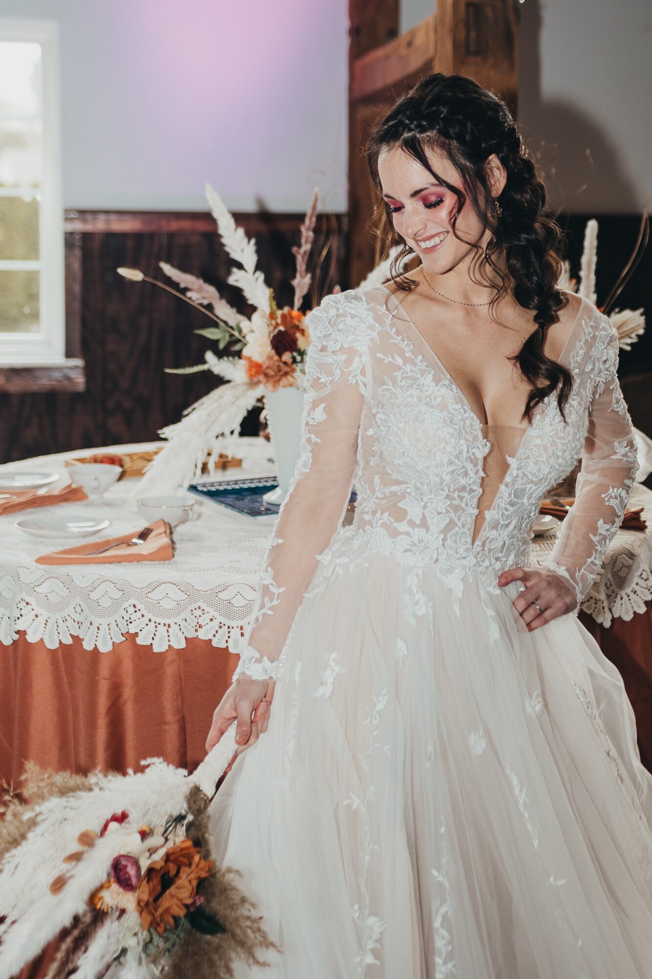 Woman in wedding dress with lace top and flowing skirt, holding bouquet, standing near decorated table, indoors.