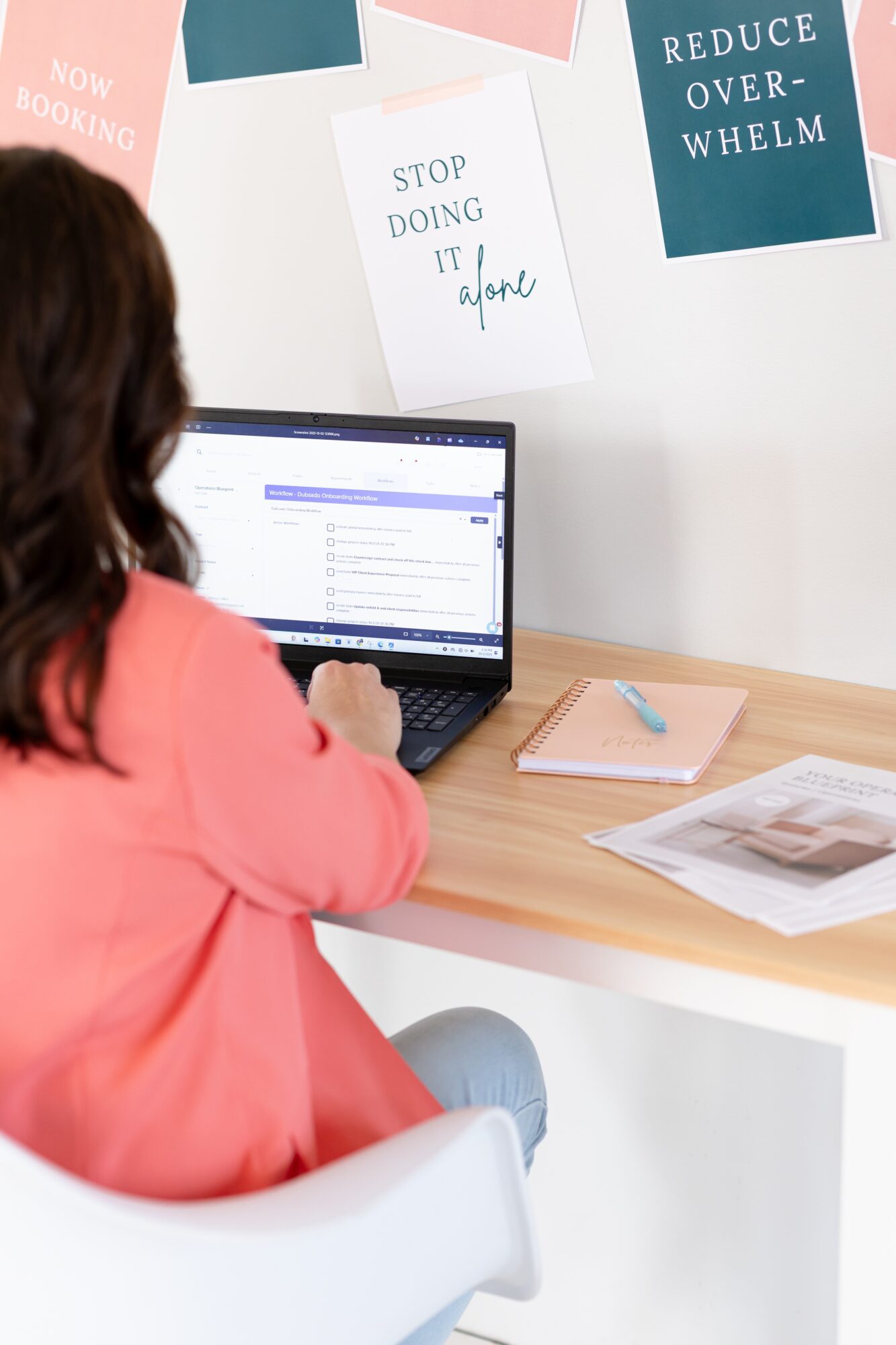 Person with dark hair in pink top working on a laptop at a desk with papers and a notebook, wall with motivational posters.