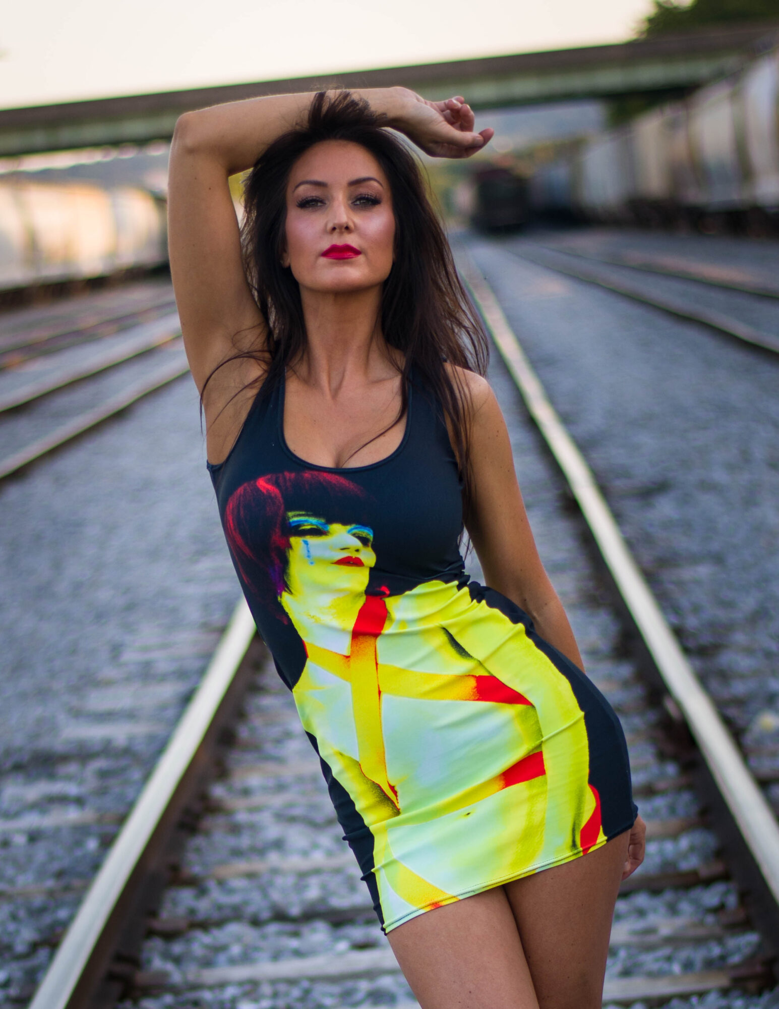 Woman with dark hair and red lipstick standing on train tracks, wearing a colorful dress, arm raised above head.
