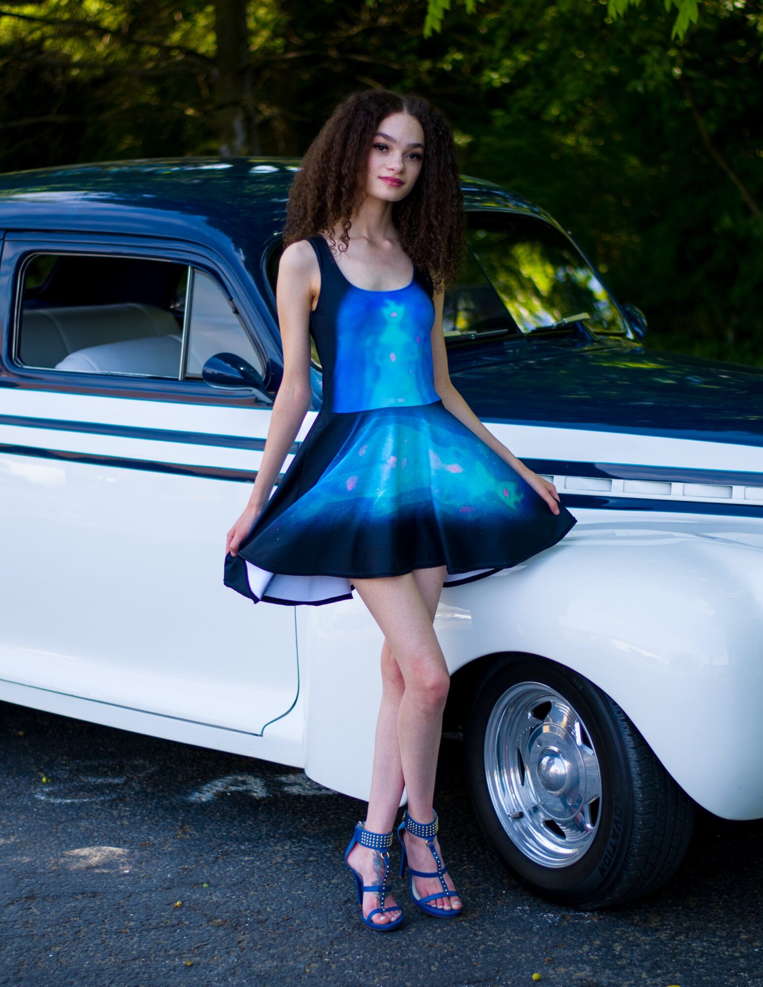 Young woman in a blue dress and high heels standing next to a vintage car outdoors, trees in background.