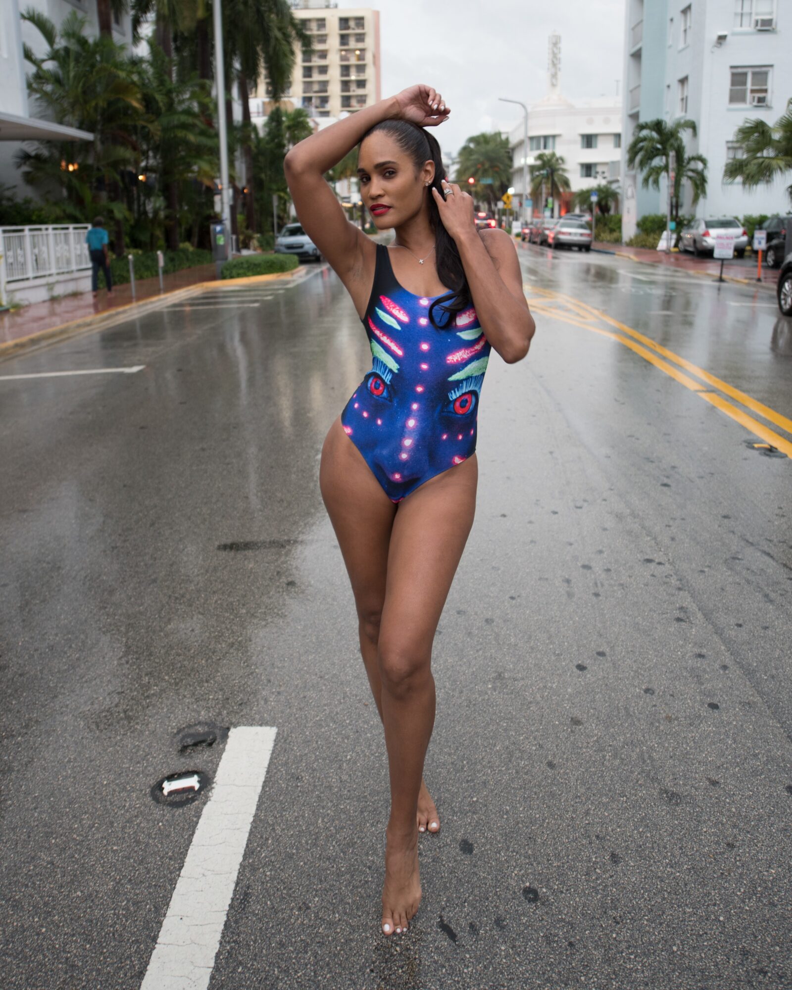 Woman in a colorful swimsuit standing on a wet city street with buildings and palm trees in the background.