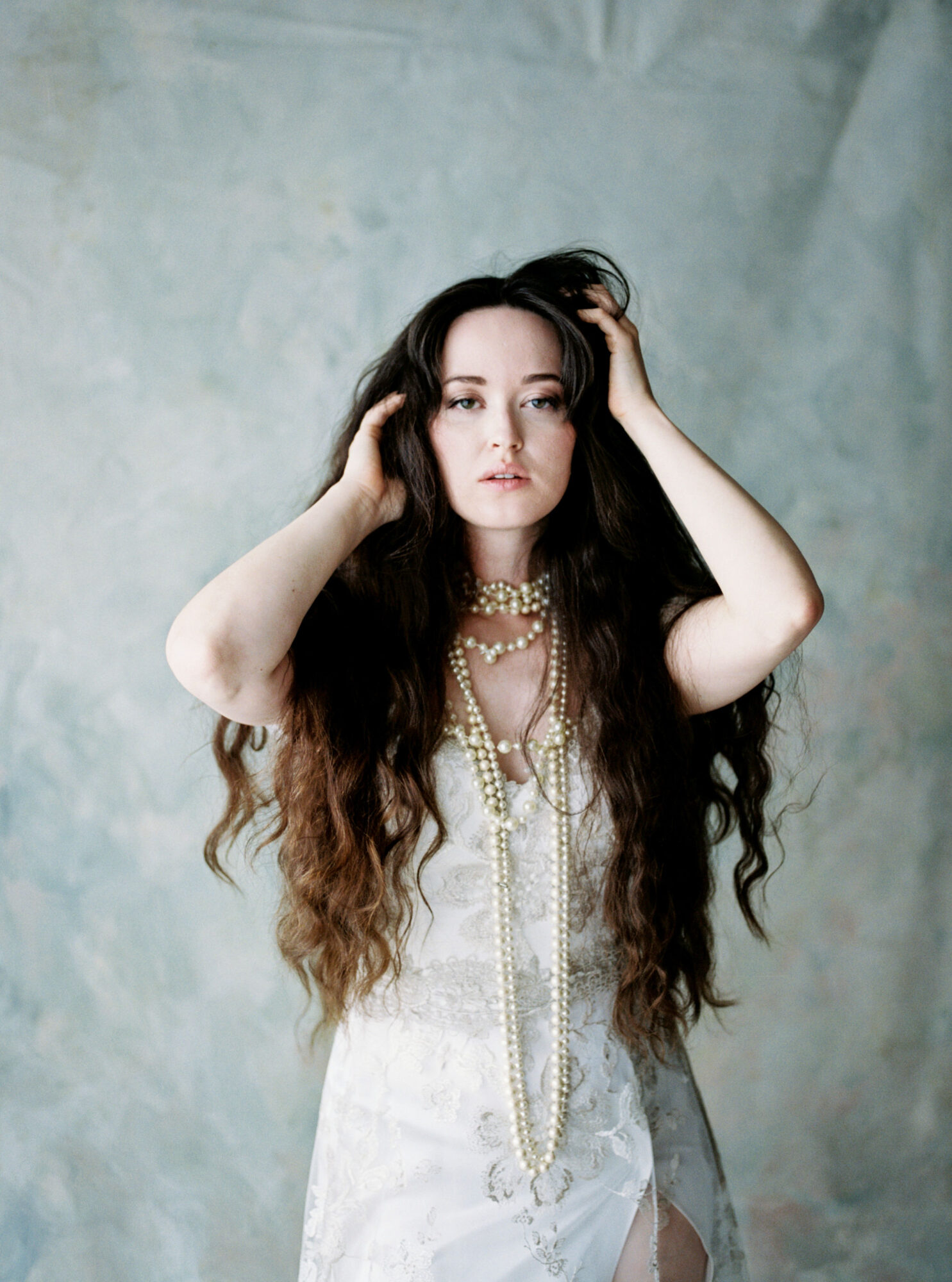 Young woman with long dark hair, wearing a white dress and layered pearl necklaces, posing with hands in hair against a textured background.