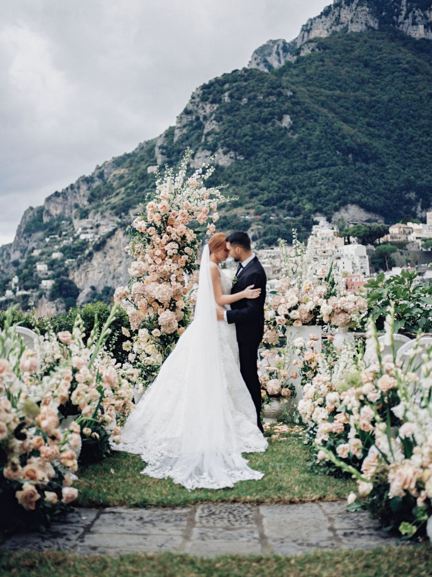 Bride and groom embrace in a garden with mountains and floral arrangements, cloudy sky in the background.