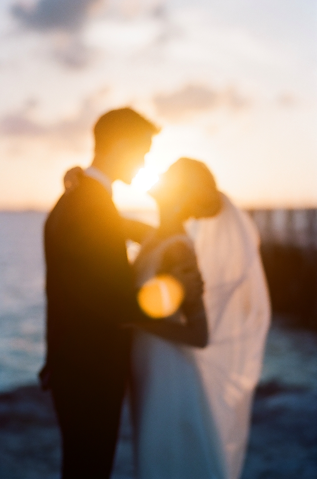 Silhouetted couple facing each other at sunset near water, with the sun behind them creating a glow.