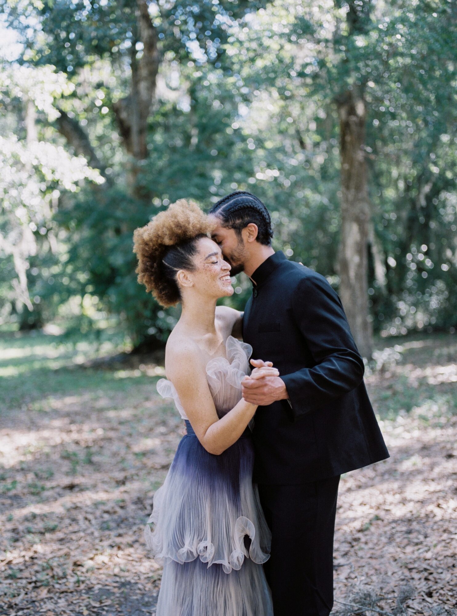 Couple dancing outdoors in a wooded area, holding hands and smiling, with trees and sunlight in the background.
