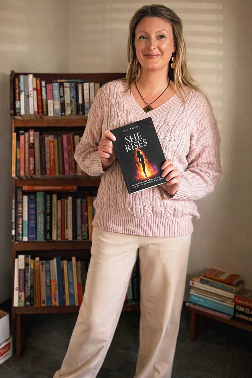 Woman holding a book titled 'She Rises' in a room with bookshelves, smiling, wearing a pink sweater and white pants.