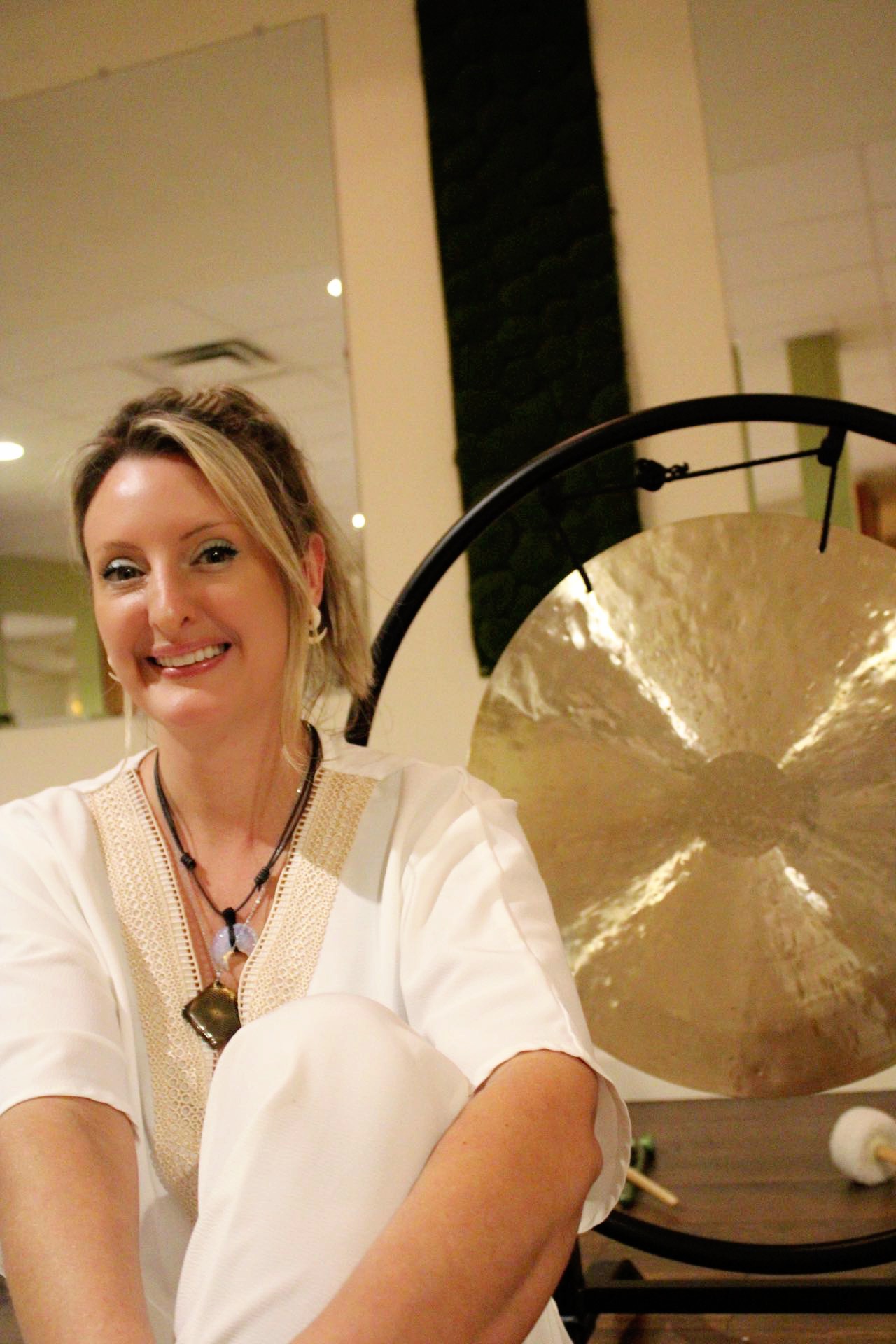 Woman smiling, sitting indoors with a large gong behind her, wearing a white top and necklace.