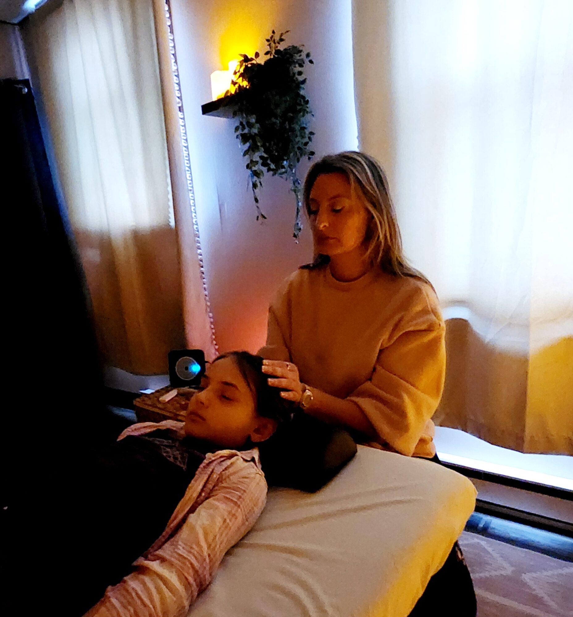 Woman gently touches child's head while lying on a bed in a softly lit room with curtains and a plant.