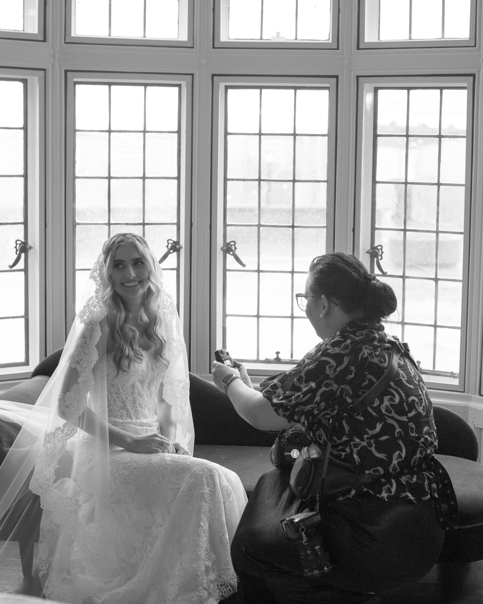 Two women sit by a large window with multiple panes, one in a wedding dress, the other taking a photo.