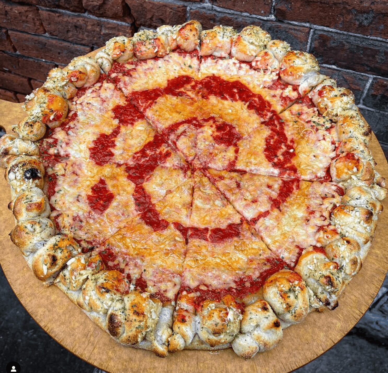 Large pizza with cheese, tomato sauce, and a crust decorated with bread rolls, on a wooden board against a brick wall.