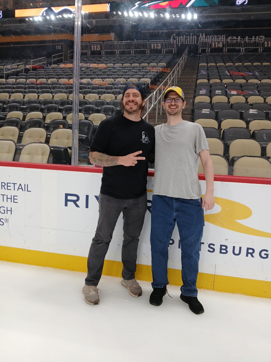 Two people stand on an ice rink, smiling, with empty stadium seats in the background.