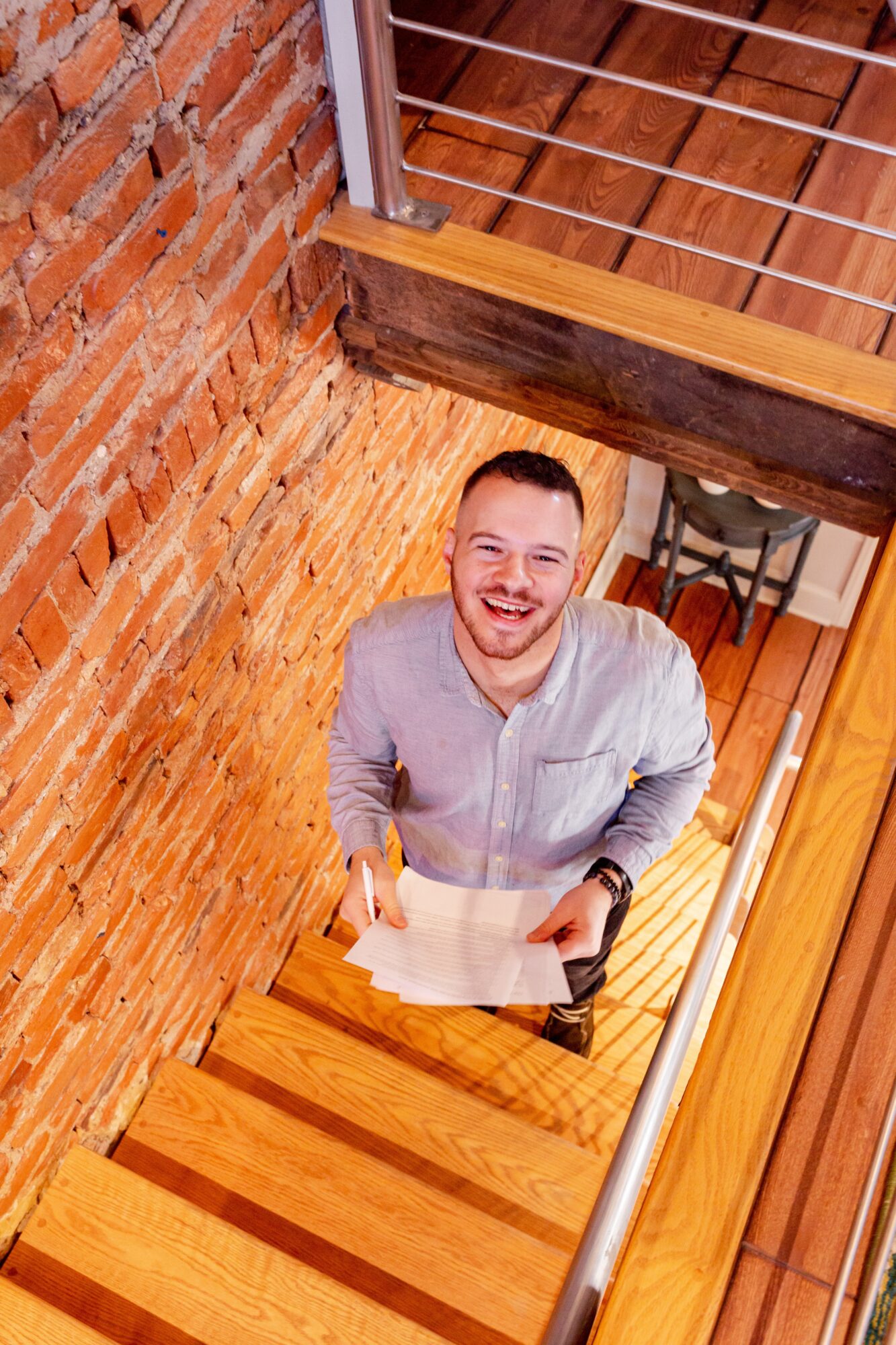 Smiling man holding papers on wooden staircase with brick wall and railing, viewed from above.