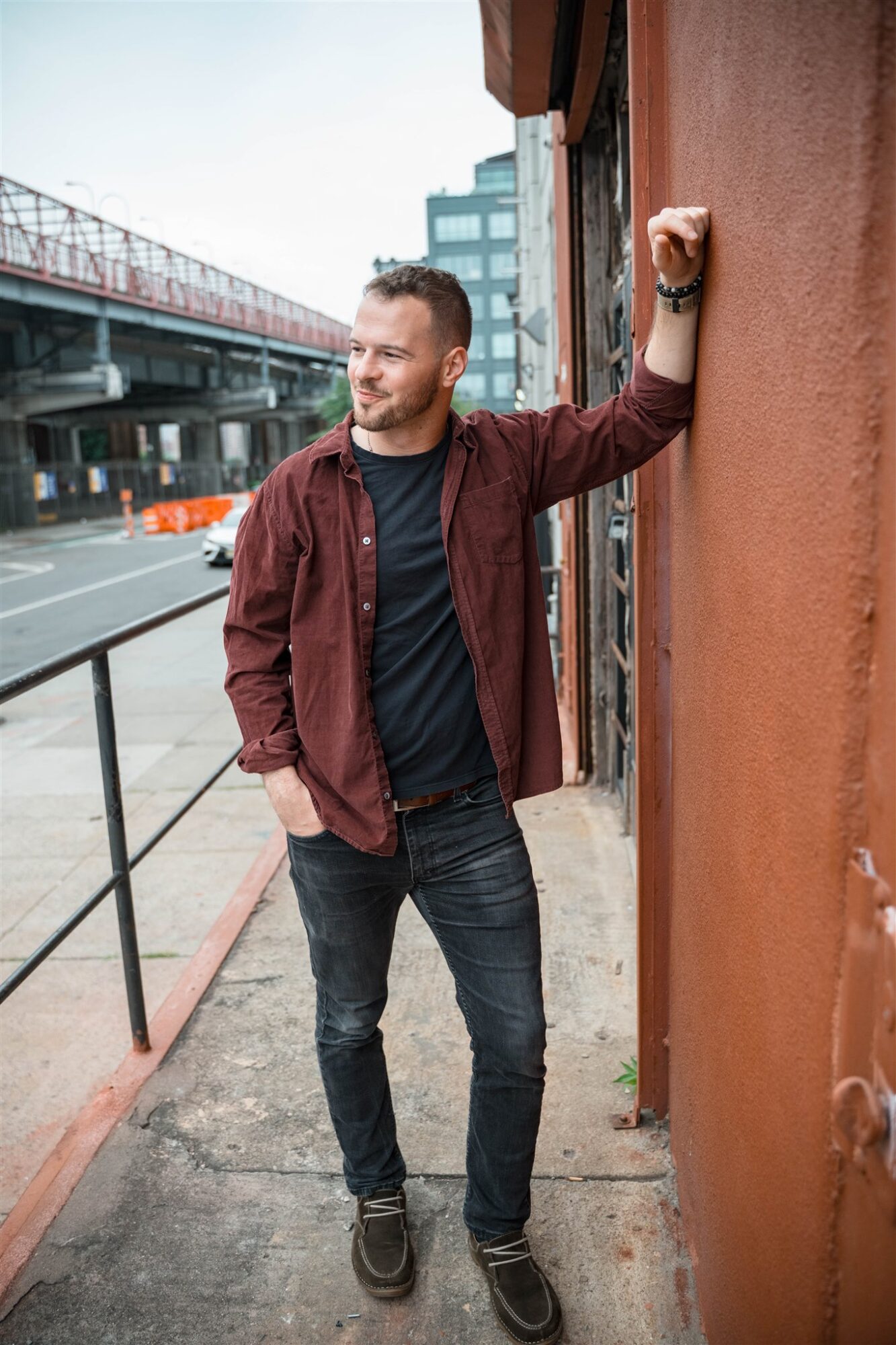 Man leaning against a wall outdoors, with city buildings and a bridge in the background.