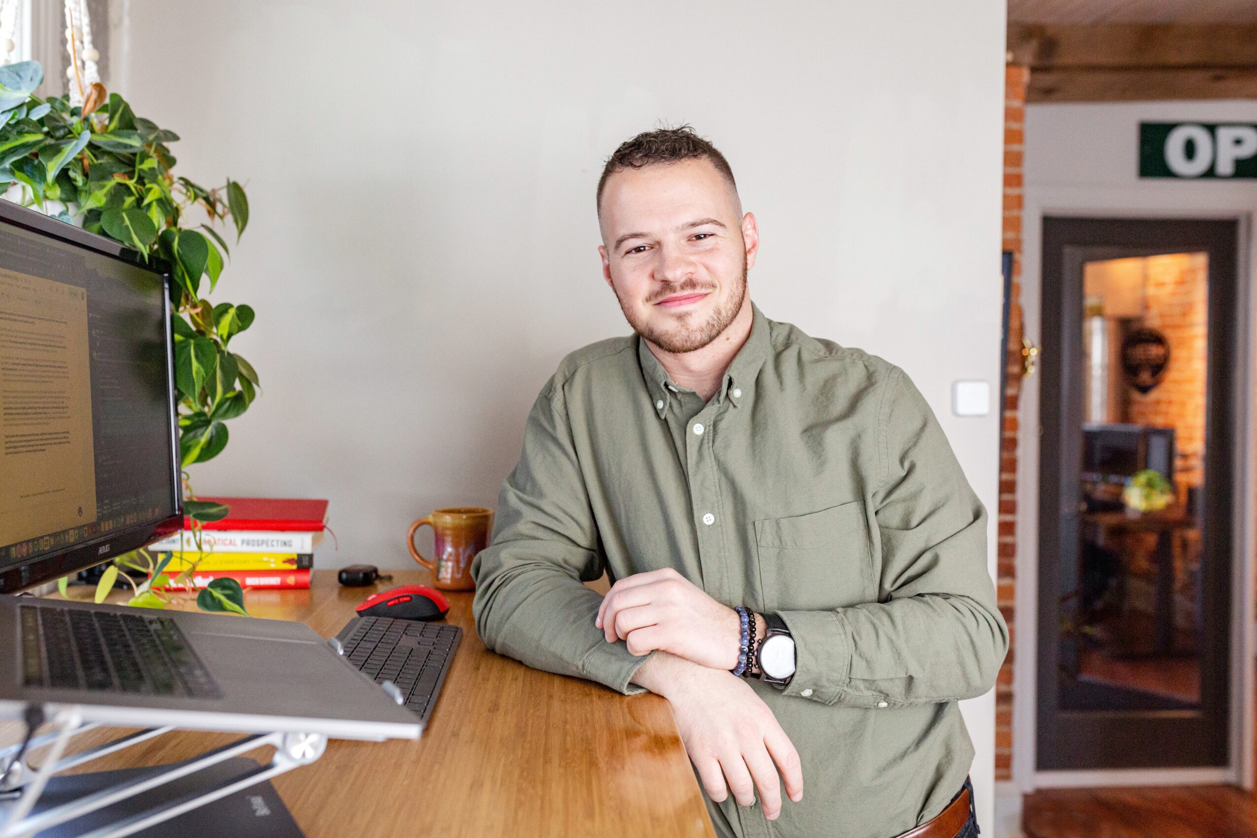 Young man with short hair and beard sitting at a desk with a computer, smiling, in a room with a plant and a door.