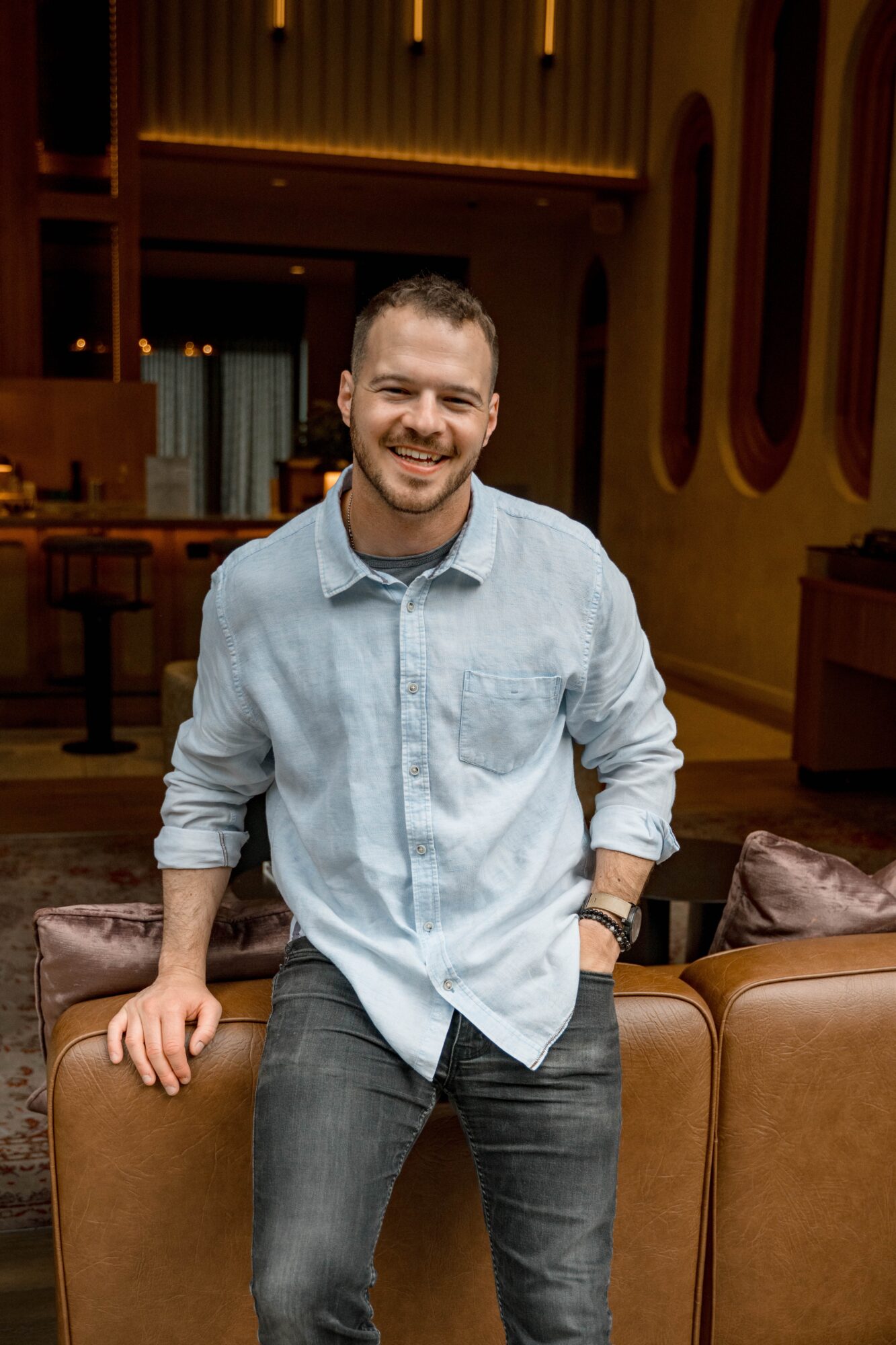 Smiling man in light blue shirt and dark jeans sitting on a brown sofa in a warmly lit room.