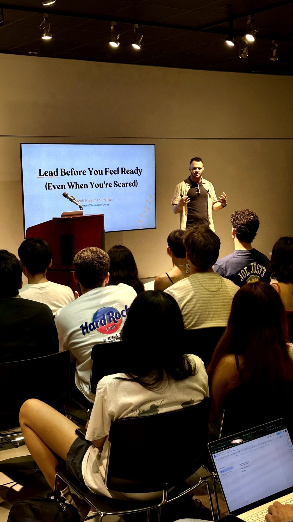 Person standing on stage giving a presentation to an audience in a room with a large screen behind them.