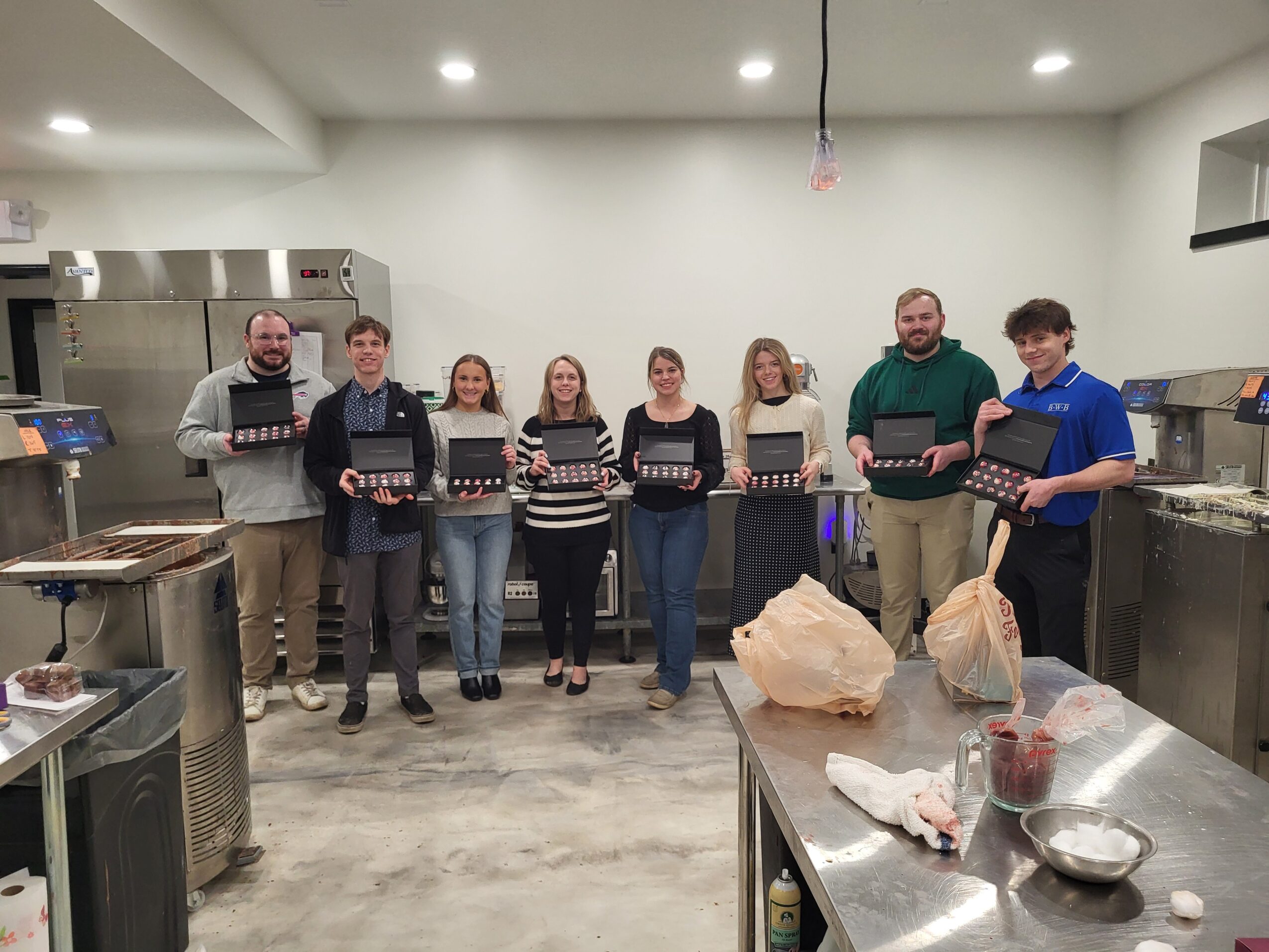 Group of eight people standing in a kitchen, holding boxes, with stainless steel appliances and a countertop with bags and bowls.