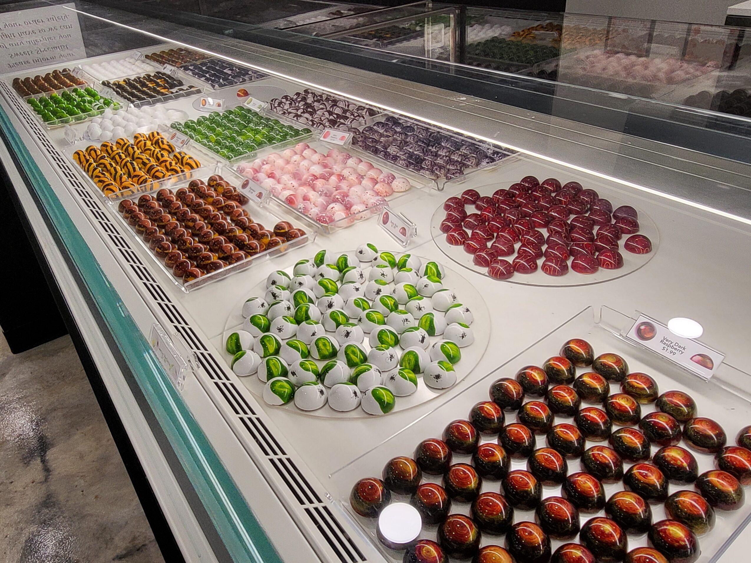 Display case with various colorful chocolates and confections arranged in rows and trays, including green and white, pink, red, and brown chocolates.