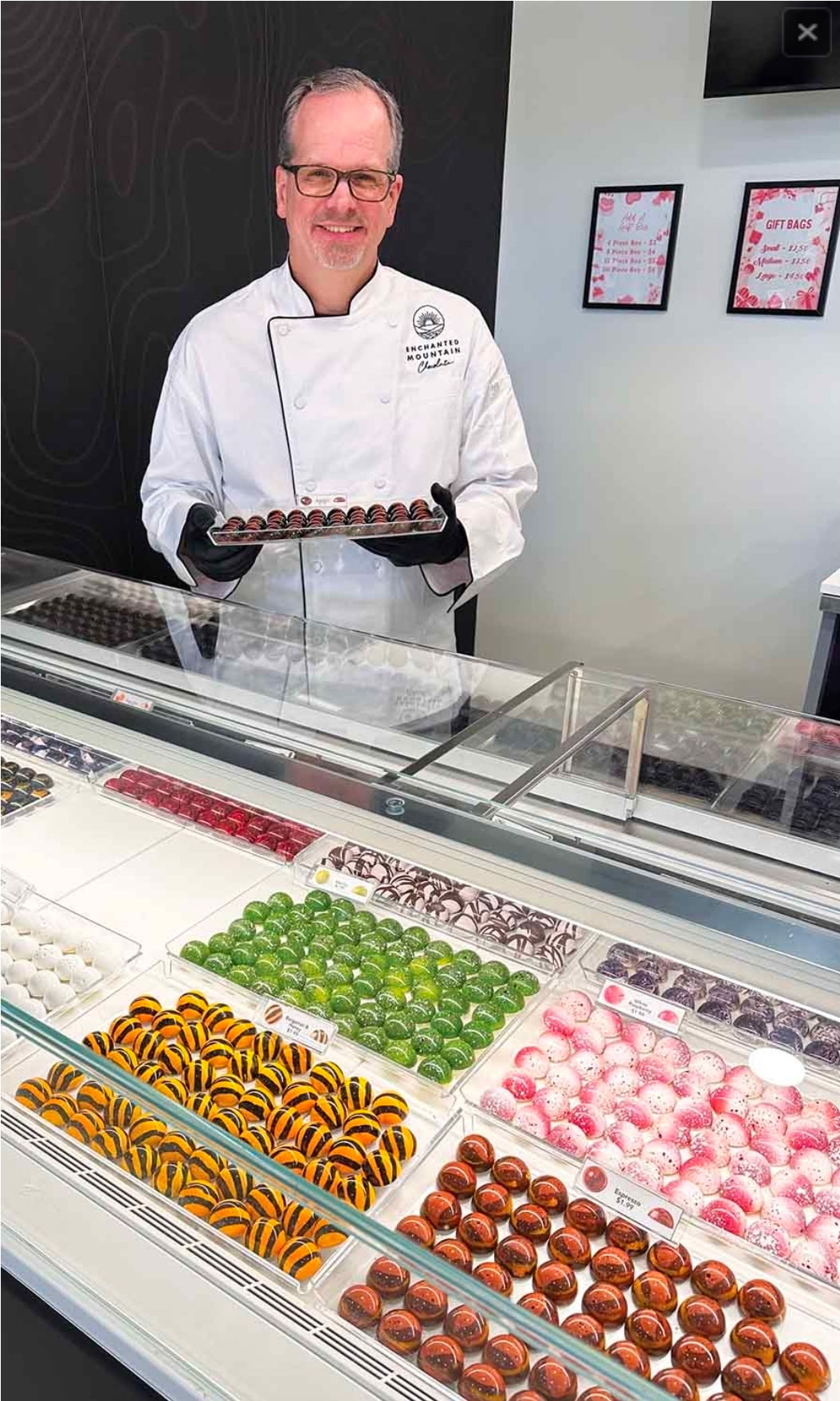 Chef in white uniform holding a tray of chocolates in a display case with colorful confections.