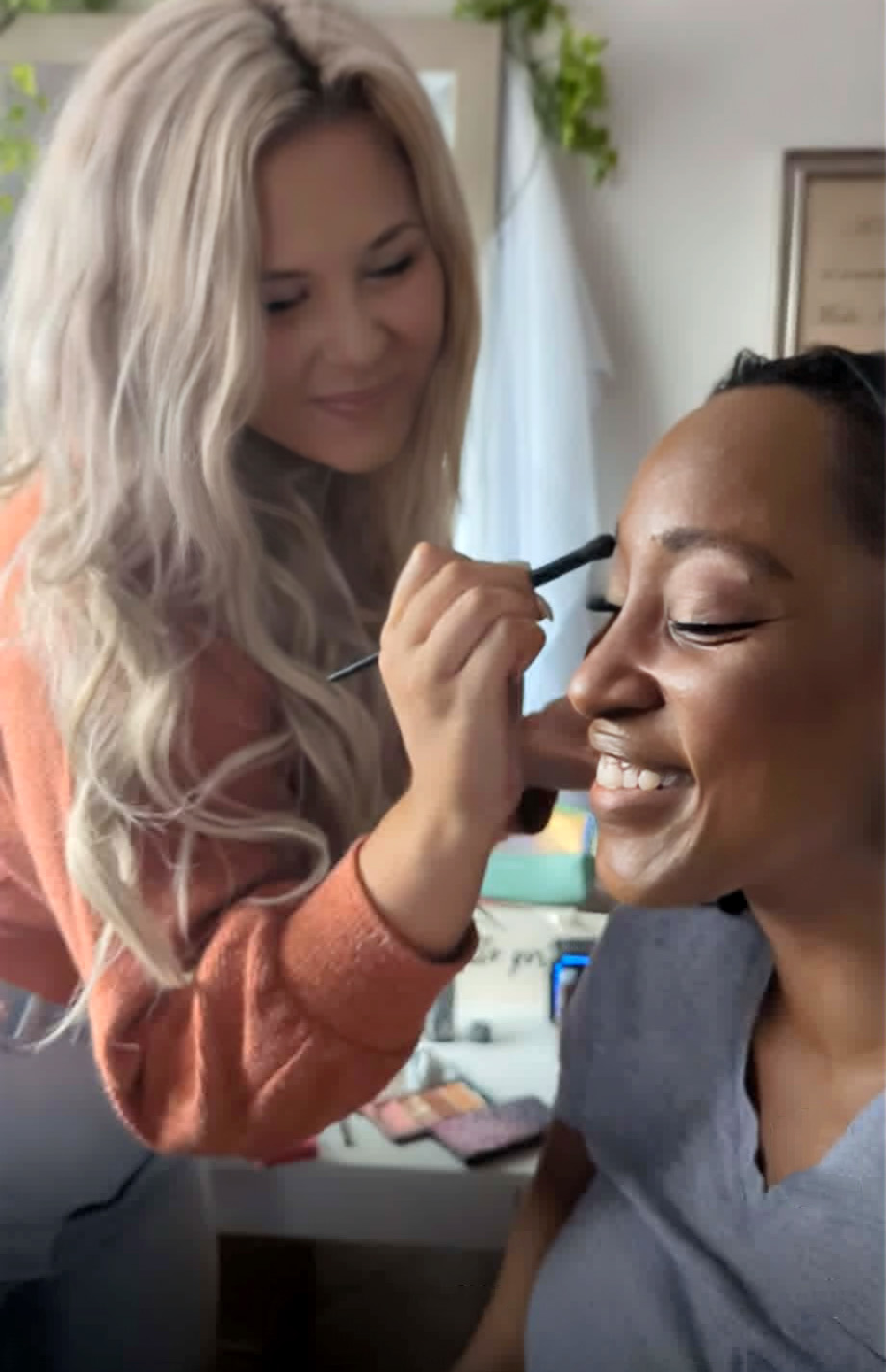 Woman applying makeup to another woman smiling, in a well-lit room with plants and framed art.