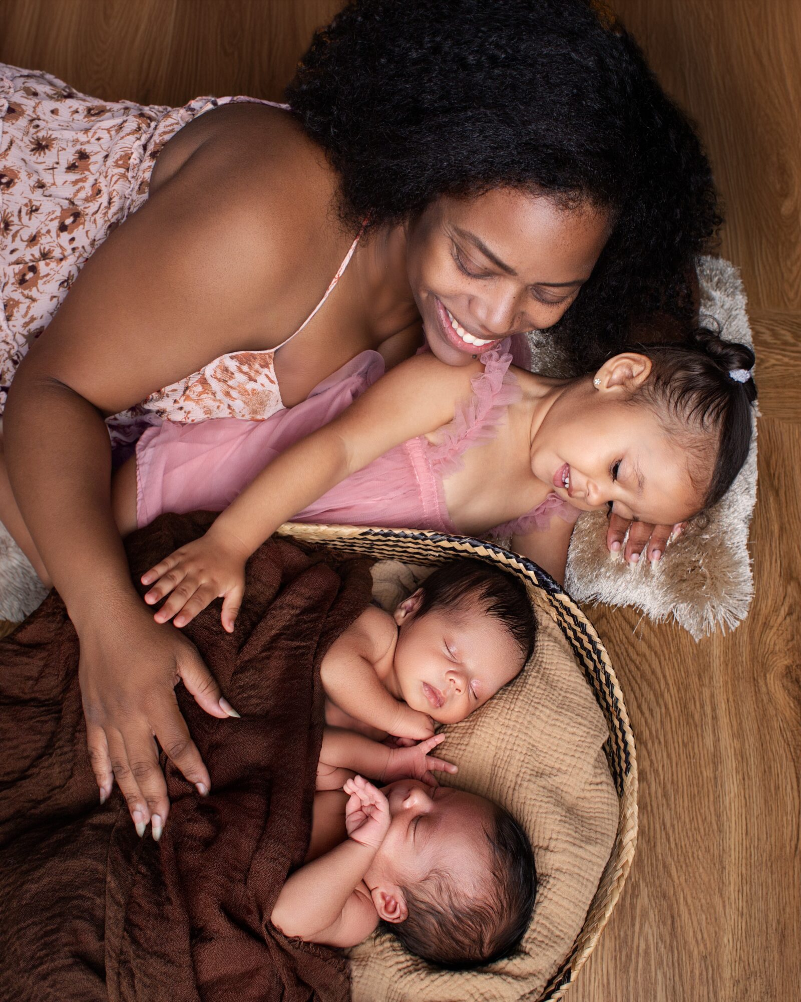 Woman and two children lying on a wooden floor, smiling and cuddling, with a basket and blanket nearby.