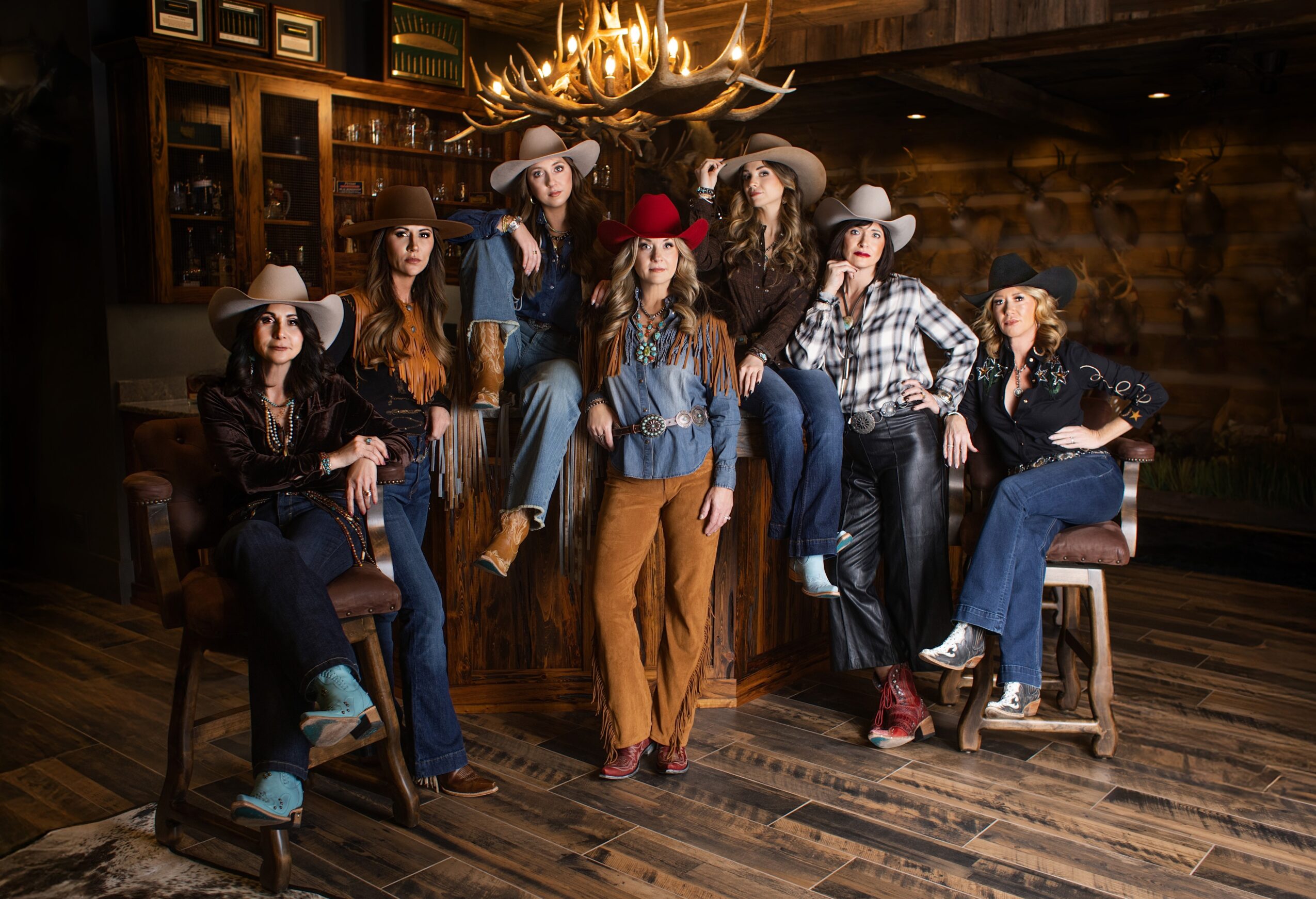 Group of women in cowboy hats and casual clothing in a rustic room with wooden walls and a chandelier.