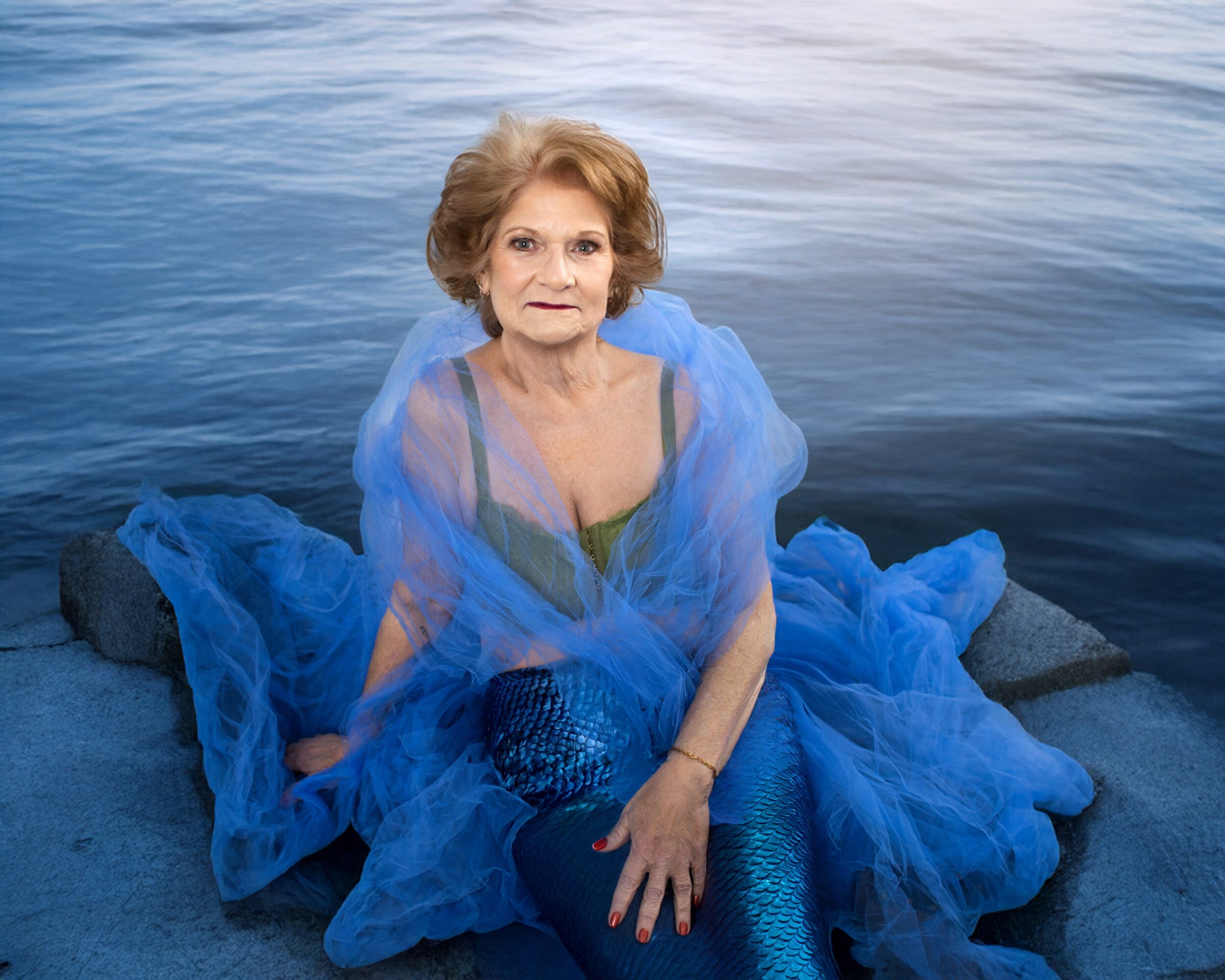 Older woman sitting on rocks by water, wearing a blue dress and sheer shawl, looking at the camera.