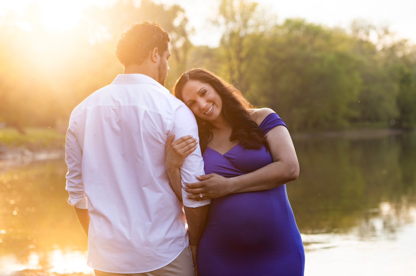 A woman in a blue dress and a man in a white shirt stand near a lake at sunset, smiling and embracing.
