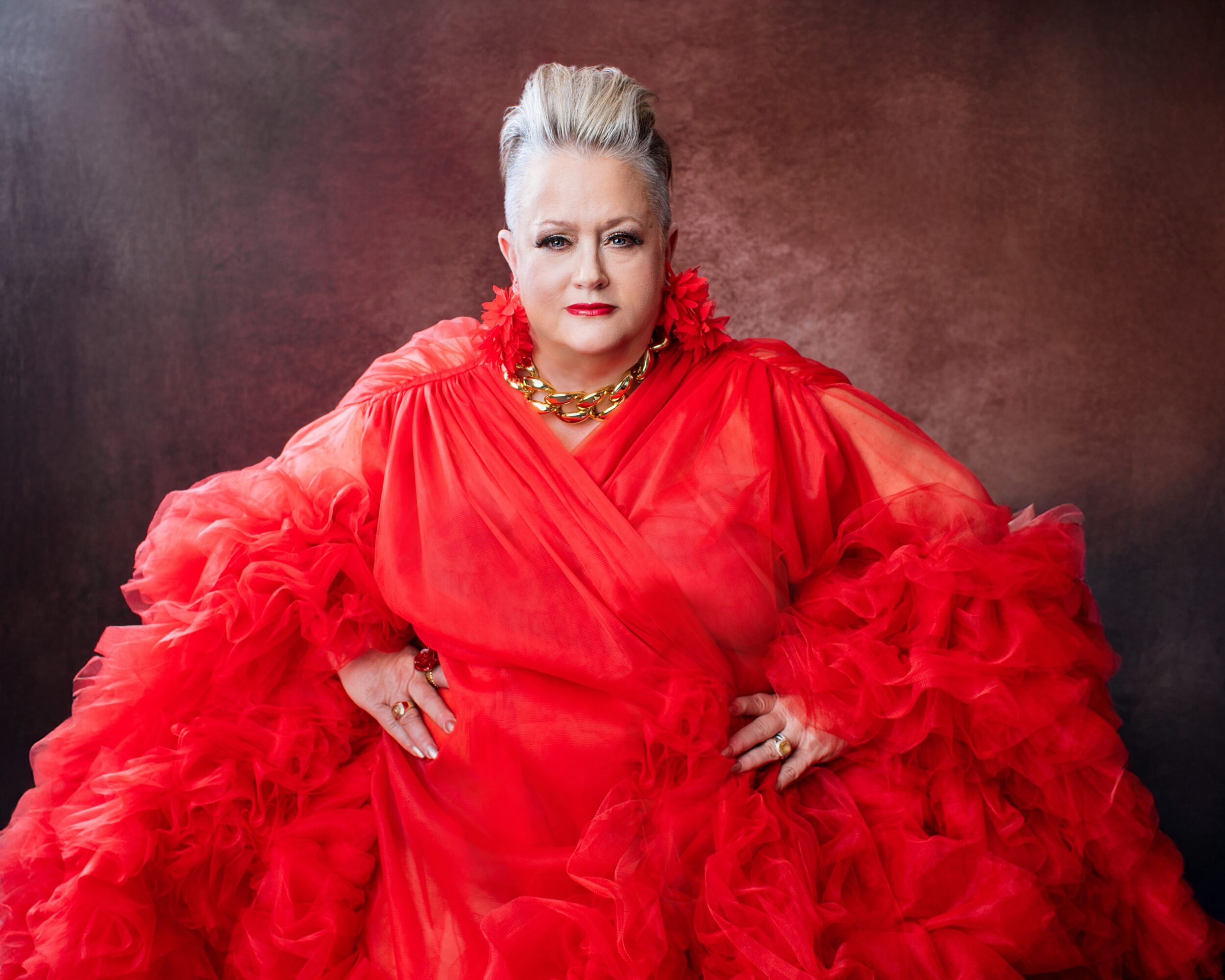 Woman in a red ruffled dress with platinum blonde hair and gold jewelry, standing against a dark background.