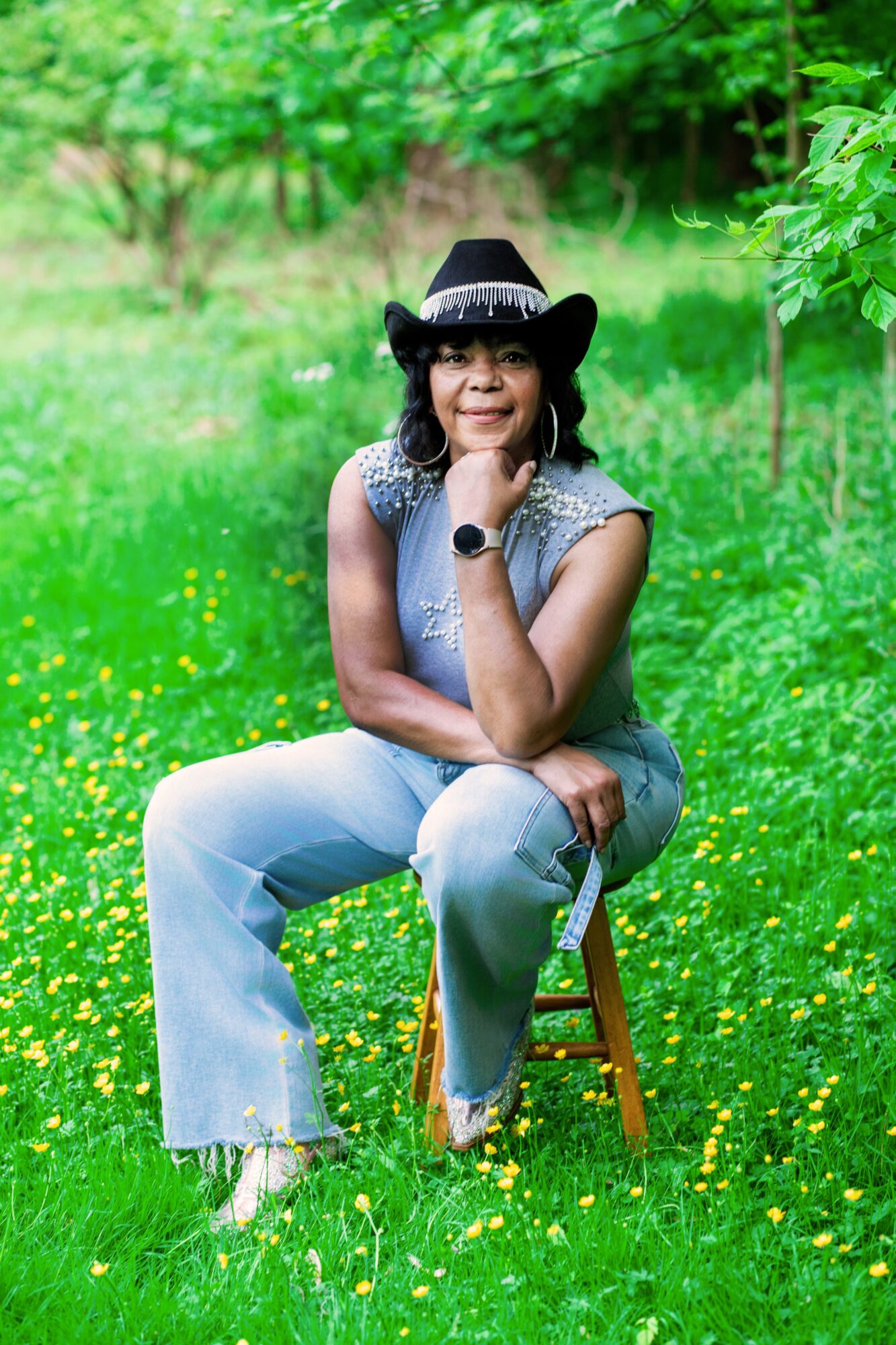 Woman sitting on a stool in a green field with yellow flowers, wearing a hat and smiling, with trees in background.