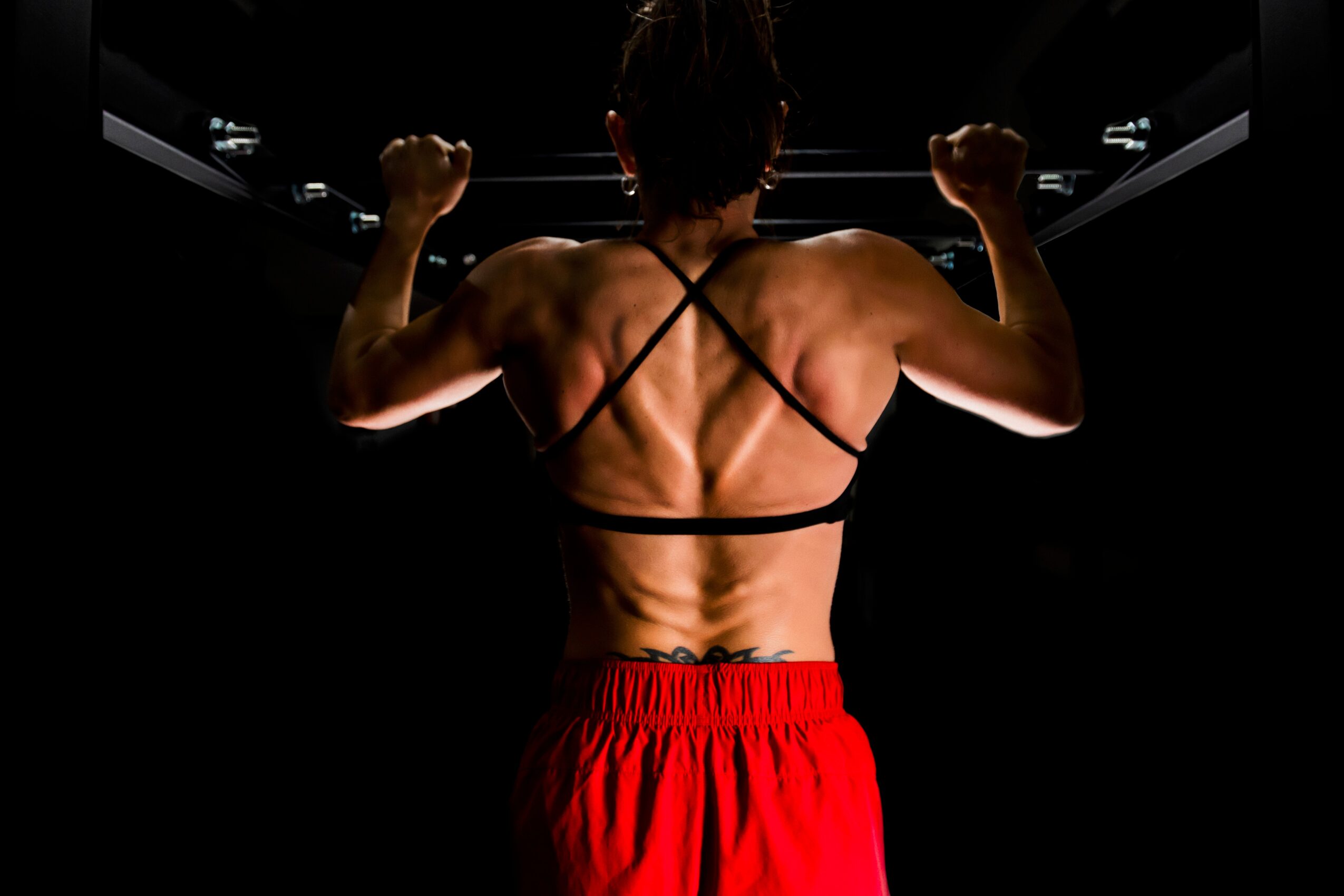 Back view of a muscular person flexing arms, wearing a strappy top and red shorts, in a dark setting.