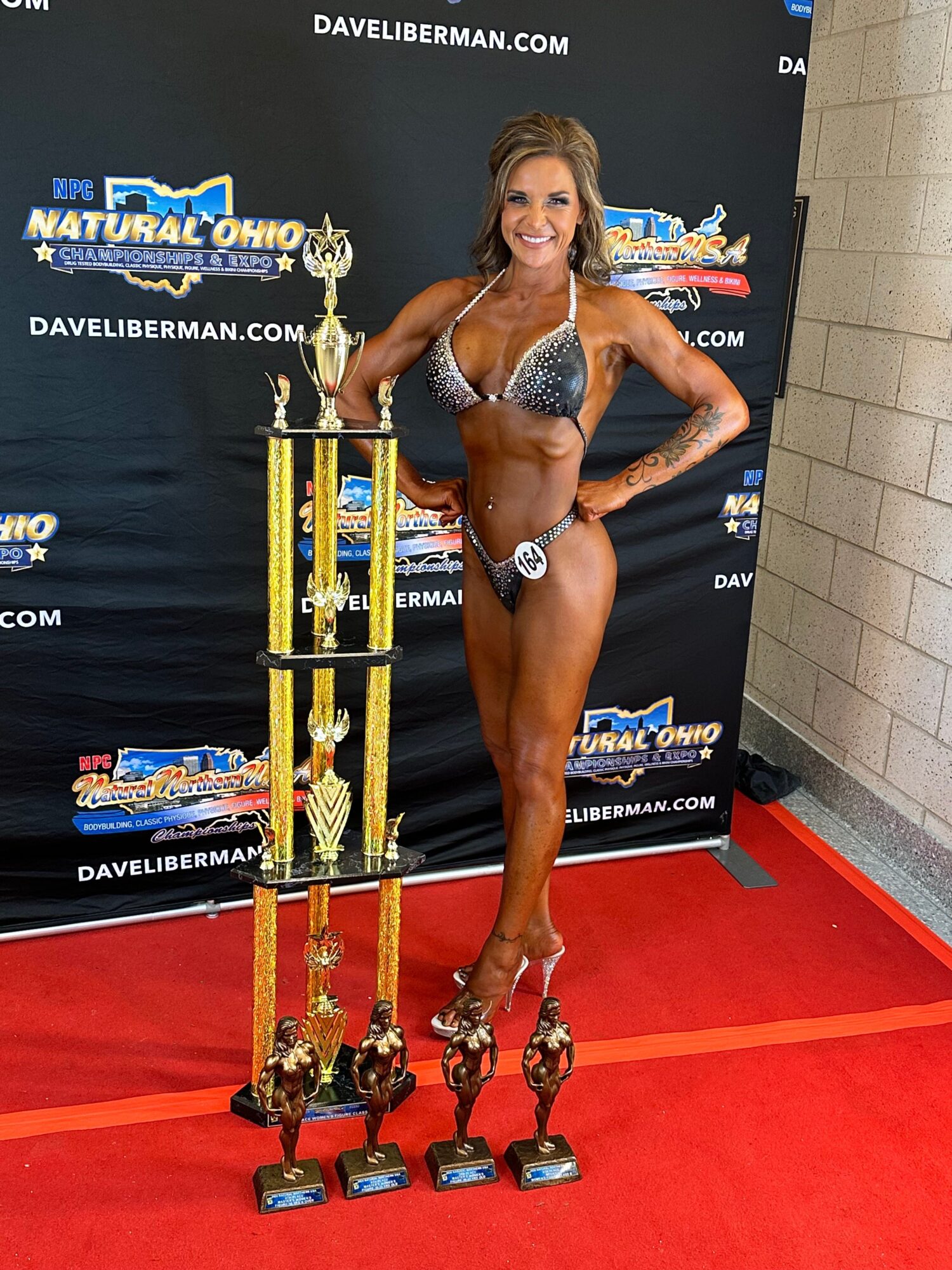 Female bodybuilder in a competition pose, wearing a silver bikini, standing on a red carpet with a large trophy in front, background with event banners.