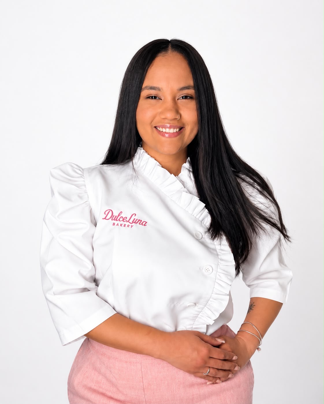 Smiling woman with long black hair wearing a white chef's coat and pink apron, standing against a white background.