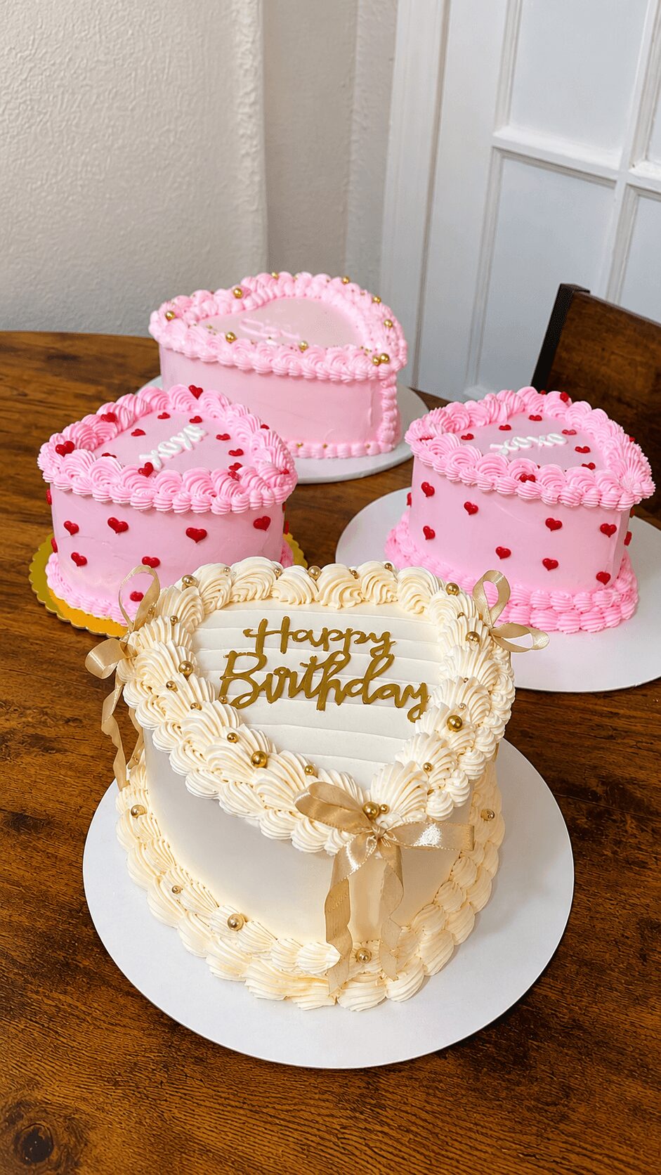 Three pink heart-shaped cakes with red and pink decorations and a white birthday cake with gold writing and cream decorations on a wooden table.