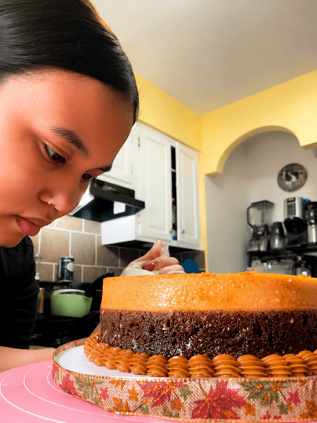 Young woman with dark hair looking at a layered cake on a pink surface in a kitchen.