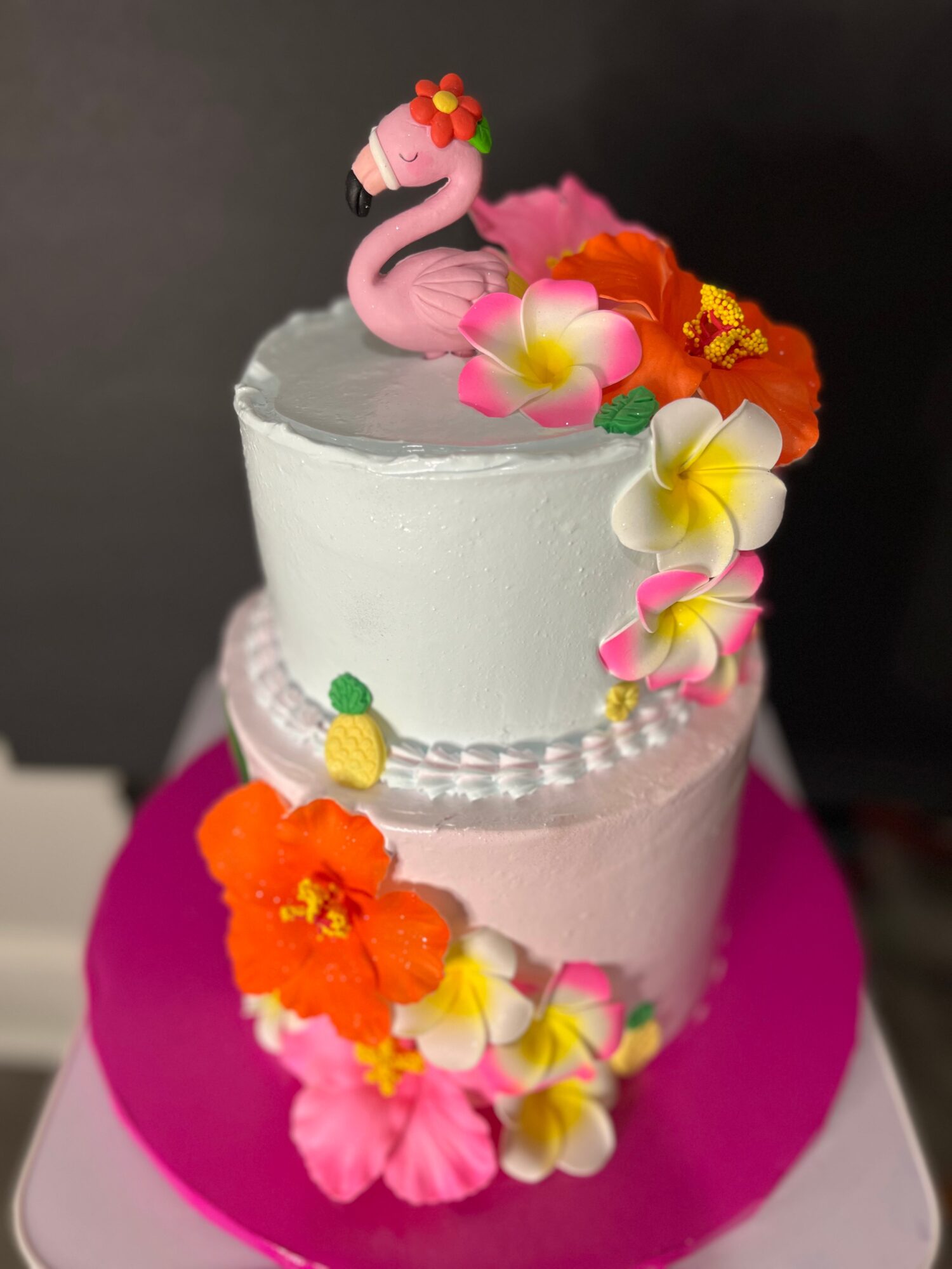 Two-tiered cake with pink and white frosting, decorated with colorful flowers and a flamingo topper, on a pink base.