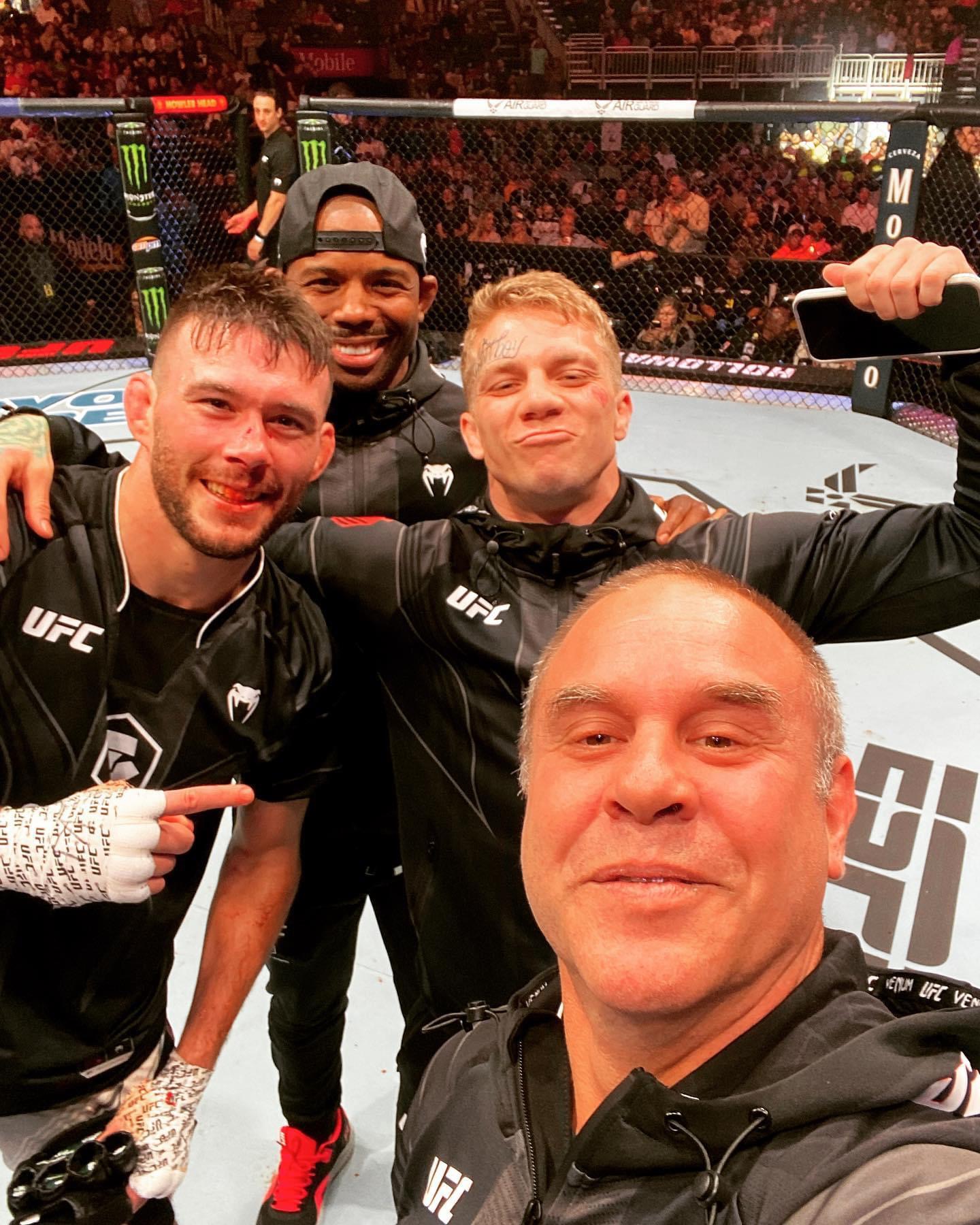 Four men in UFC gear inside an octagon, smiling and celebrating after a fight, with a crowd in the background.
