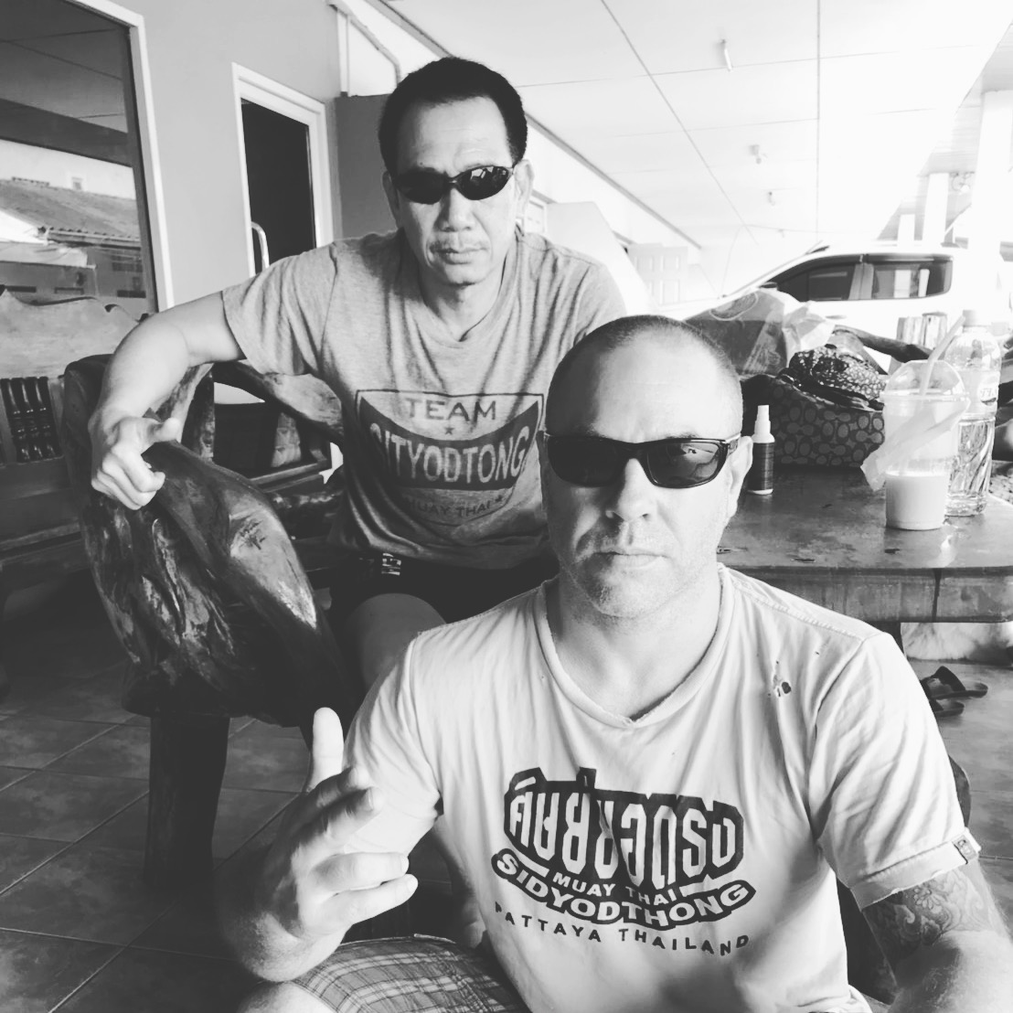 Two men wearing sunglasses sit at a table outdoors, one holding a large fish, both looking at the camera.