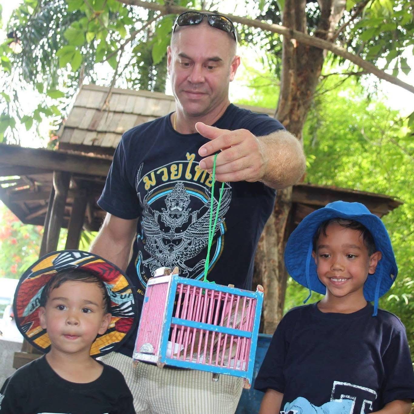 Man and two children outdoors, holding a pink birdcage, smiling, wearing hats, surrounded by trees and greenery.