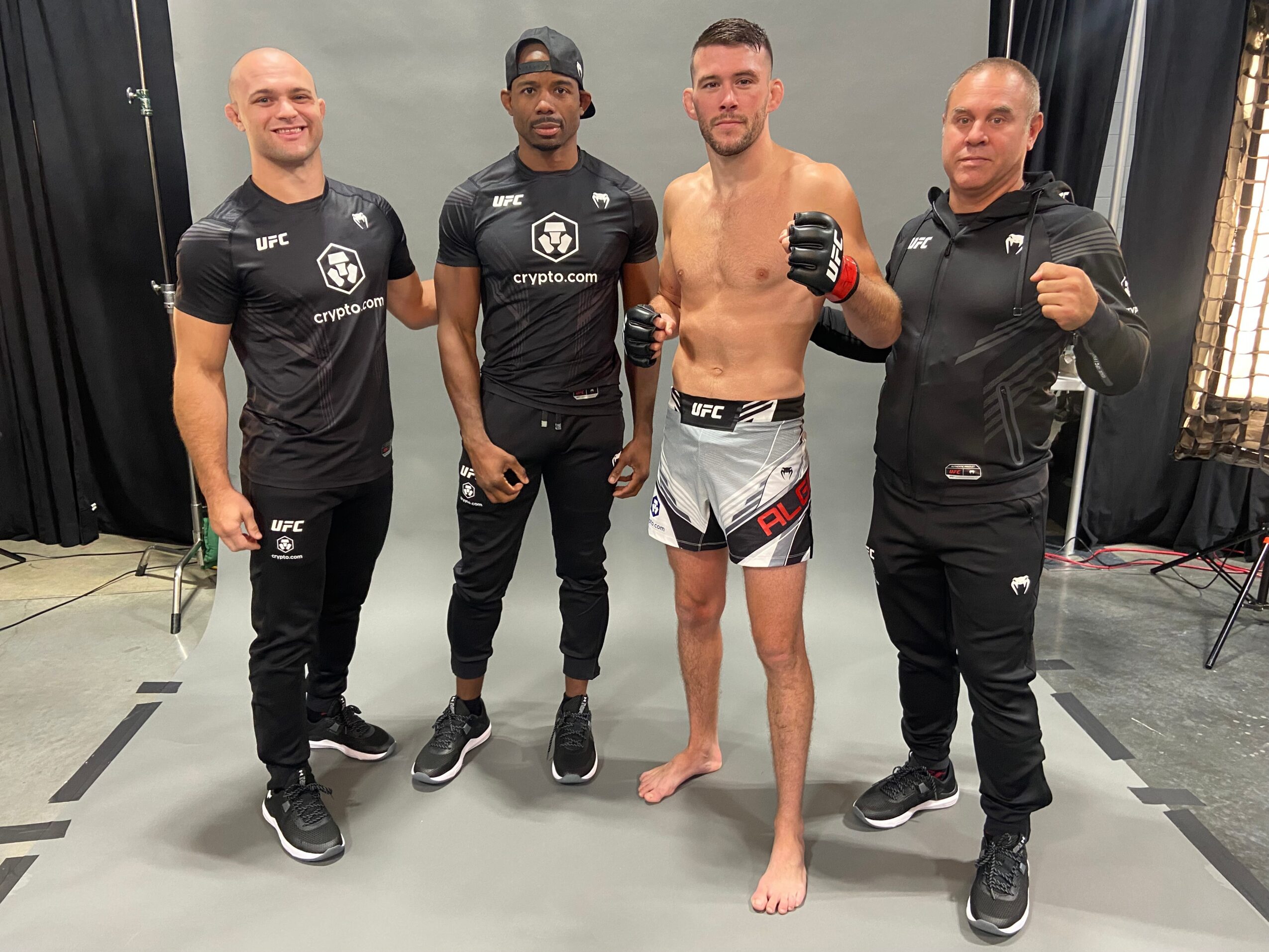 Four men standing on a mat, one shirtless with gloves, others in athletic clothing, in an indoor setting.